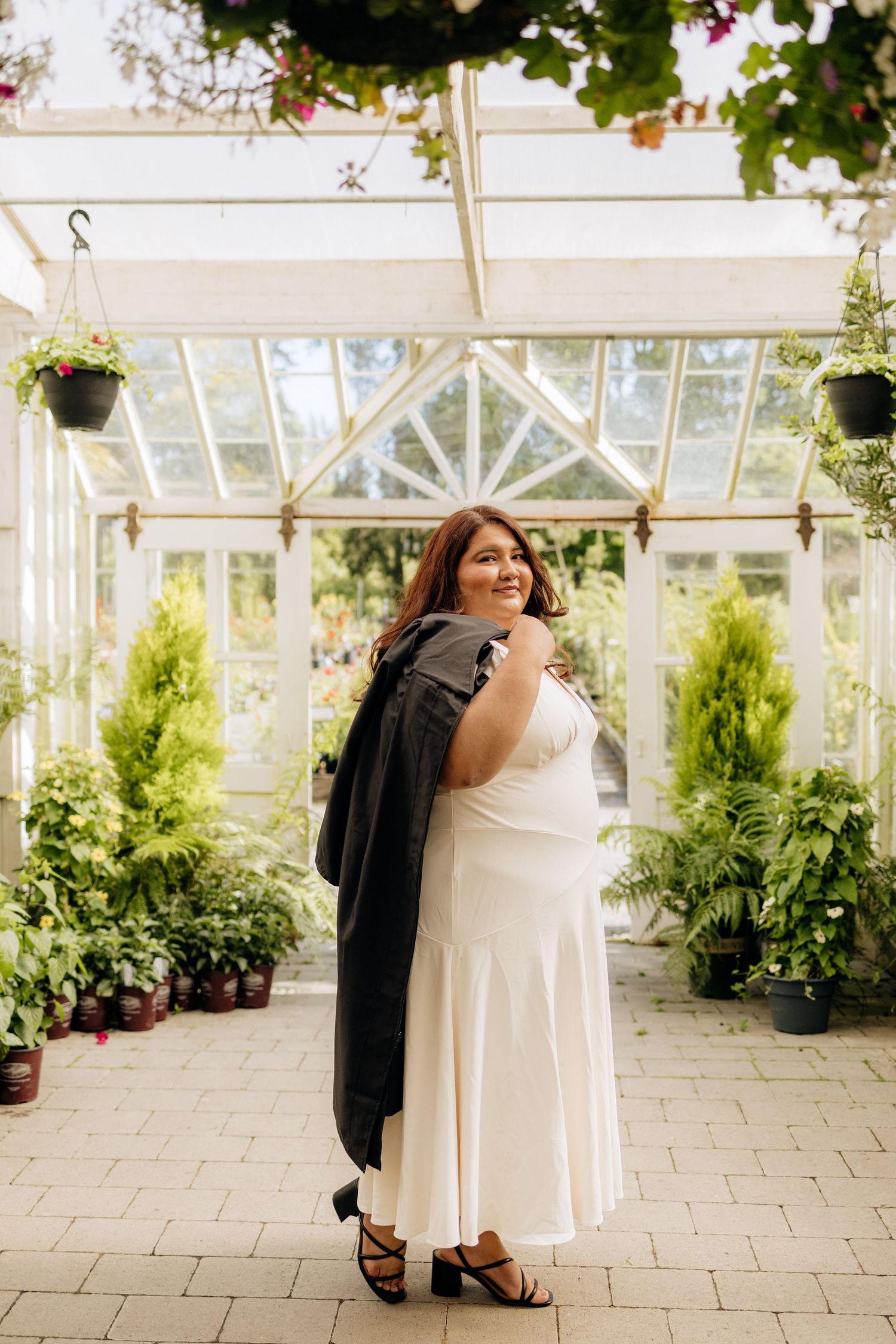 A graduate in a white dress and black jacket poses in a greenhouse surrounded by lush plants and flowers.