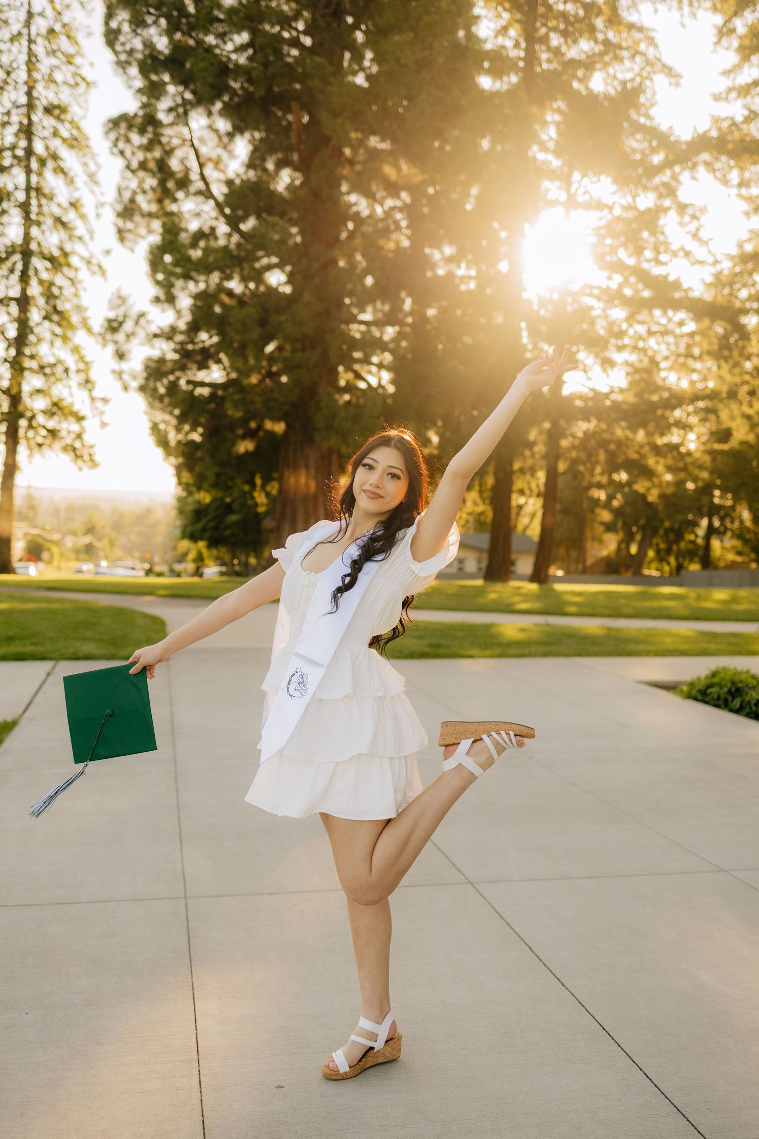 A graduate in a white dress strikes a joyful dancing pose while holding a book on a sunny campus sidewalk at sunset.