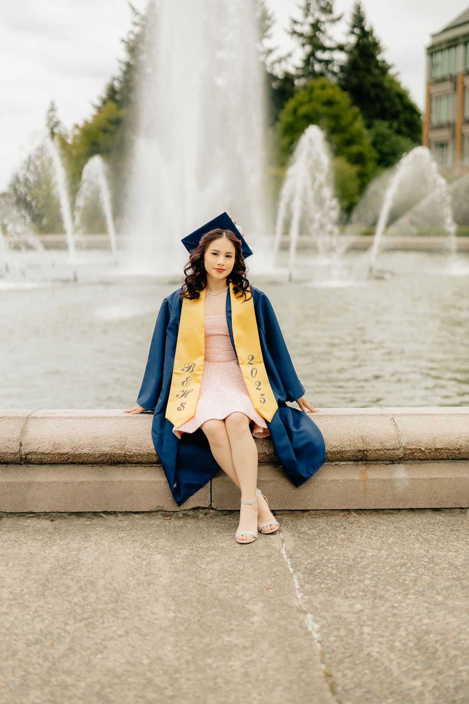 A graduate in blue and gold regalia sits by a fountain wearing a pink dress on a graduation day celebration.