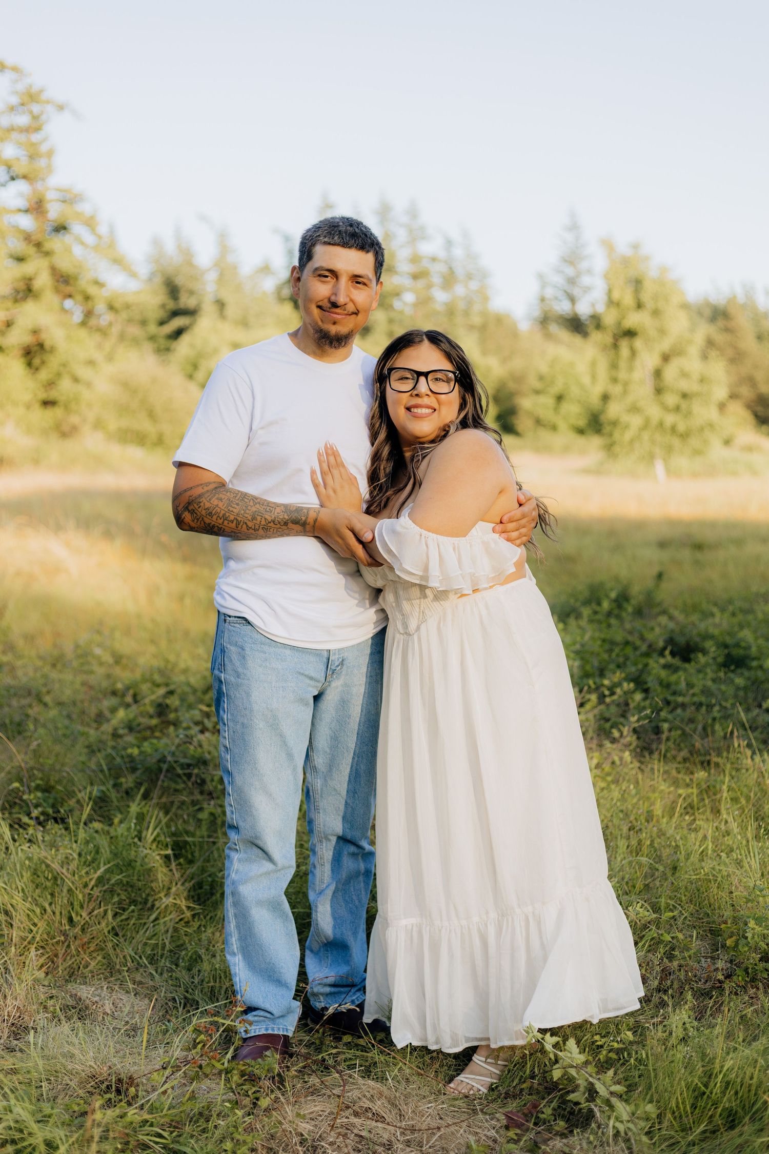 A couple embraces outdoors in a sunlit field, wearing casual white clothing against a backdrop of evergreen trees.