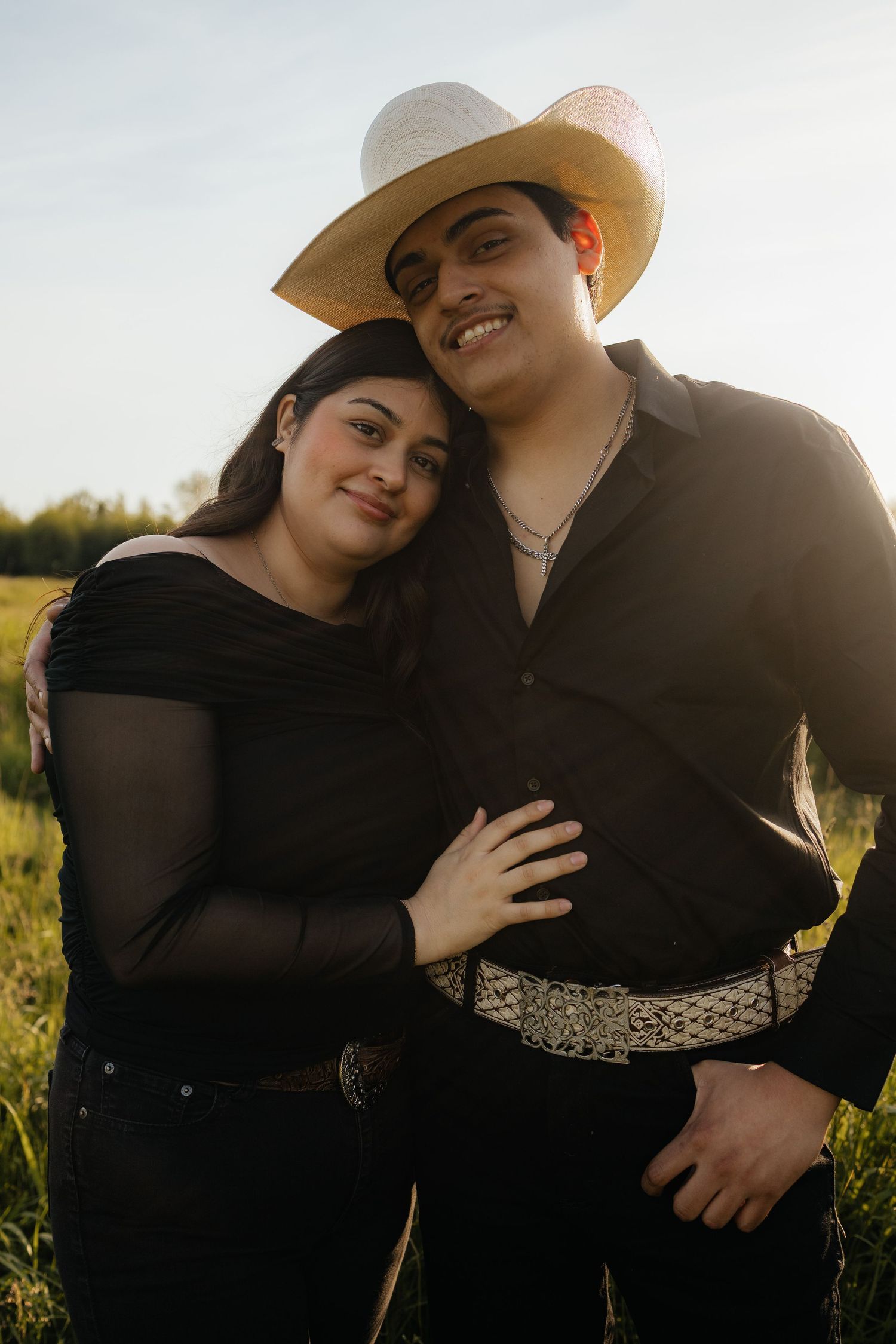 A couple wearing matching black outfits and western attire embrace in a sunlit field during golden hour.