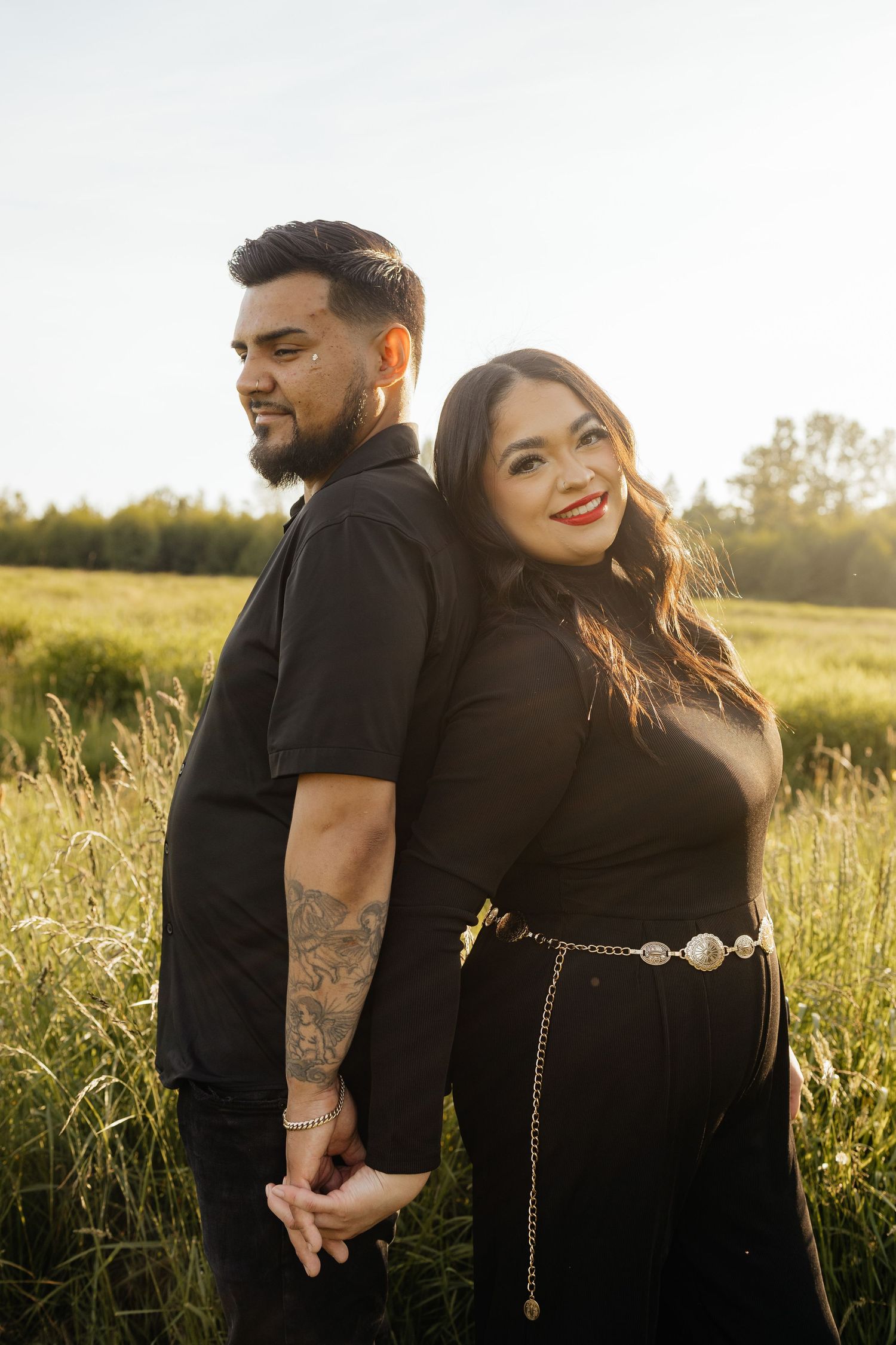 A couple in black outfits share romantic moments together in a sunlit grassy field at golden hour.