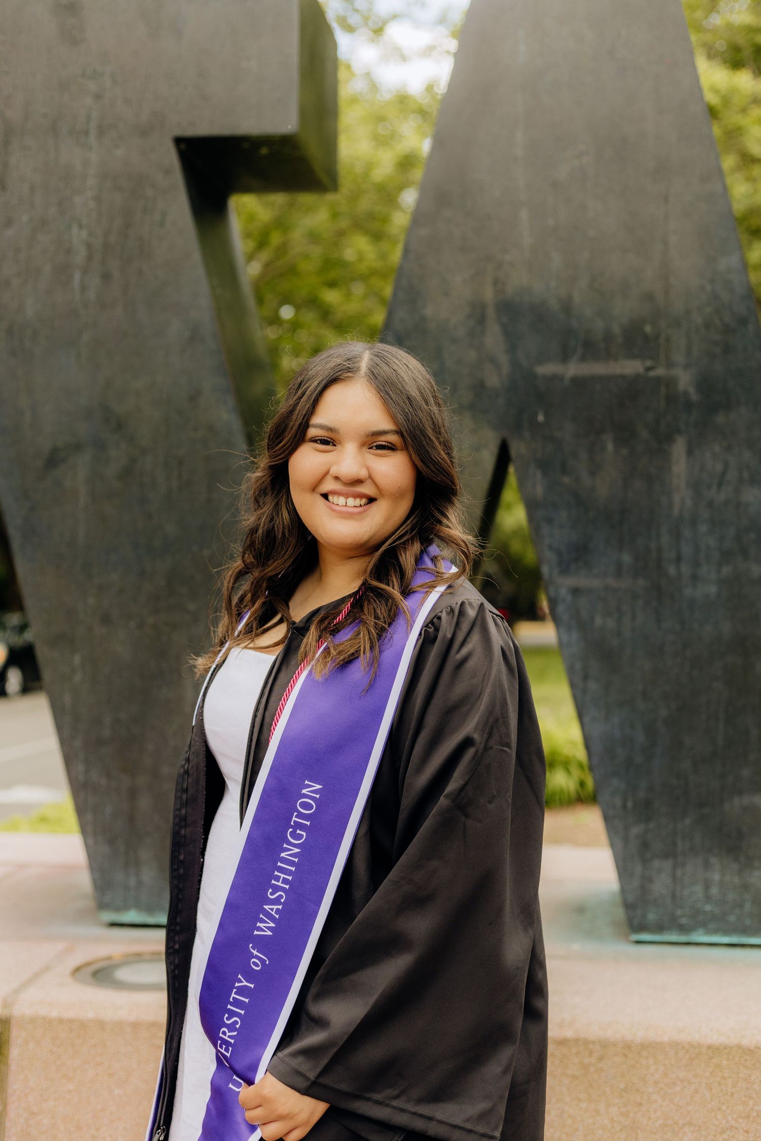 Graduate stands wearing a purple academic sash and black graduation gown next to a modern sculpture outdoors.