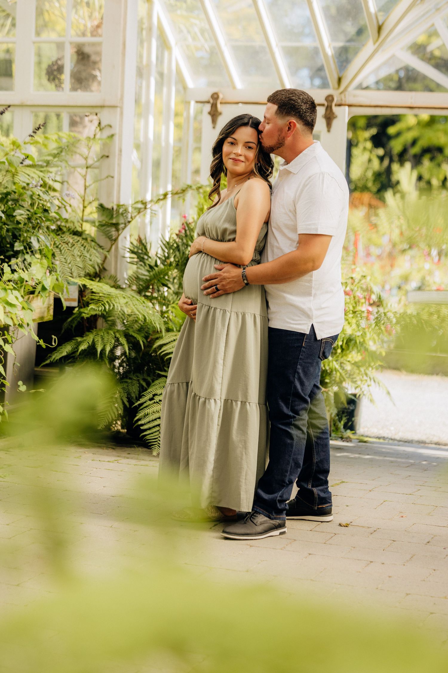A couple embraces in a romantic pose inside a bright greenhouse filled with lush green plants and natural light.