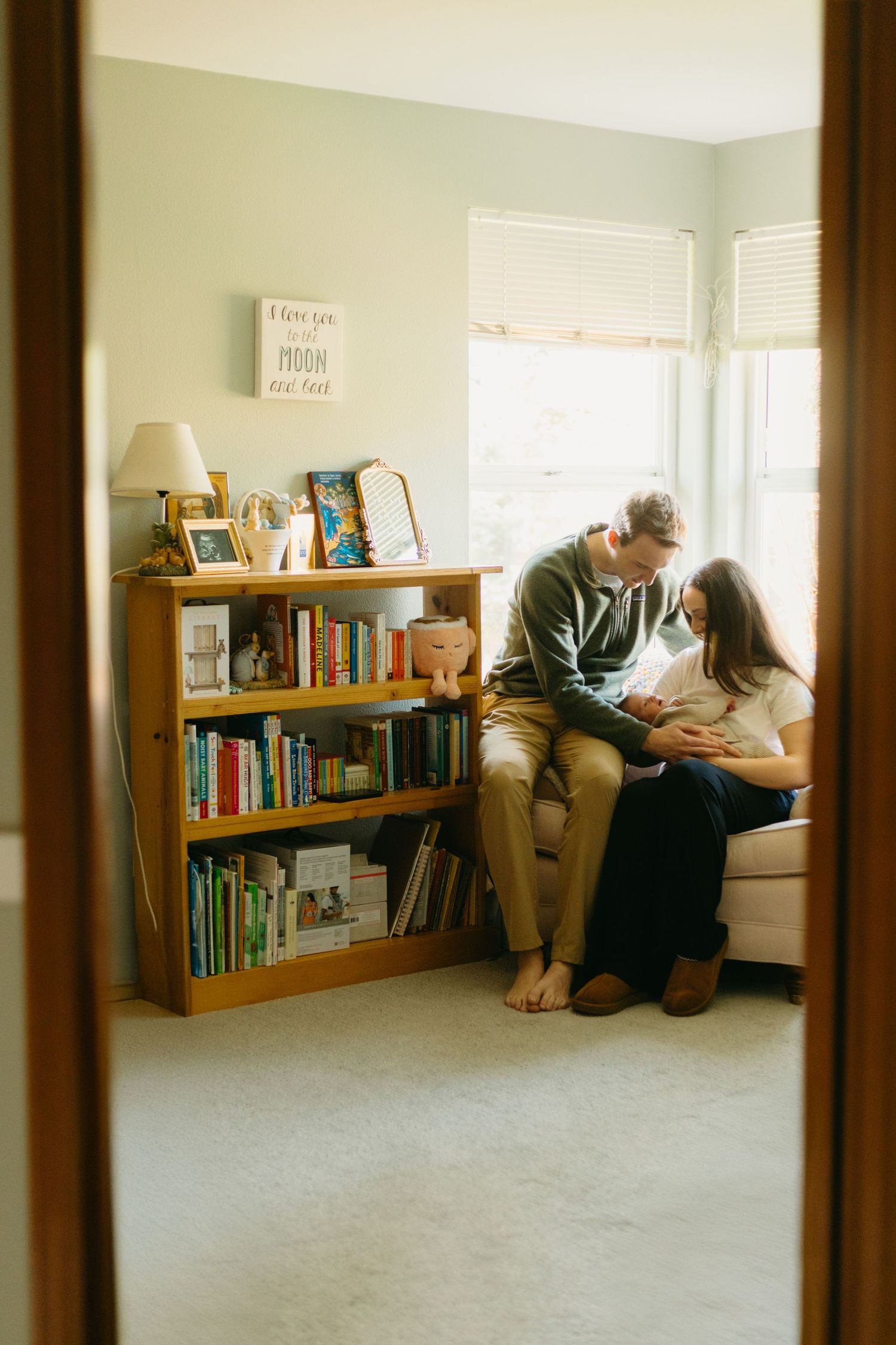 A cozy reading nook features a wooden bookshelf and window seat where a couple shares an intimate moment in warm sunlight.