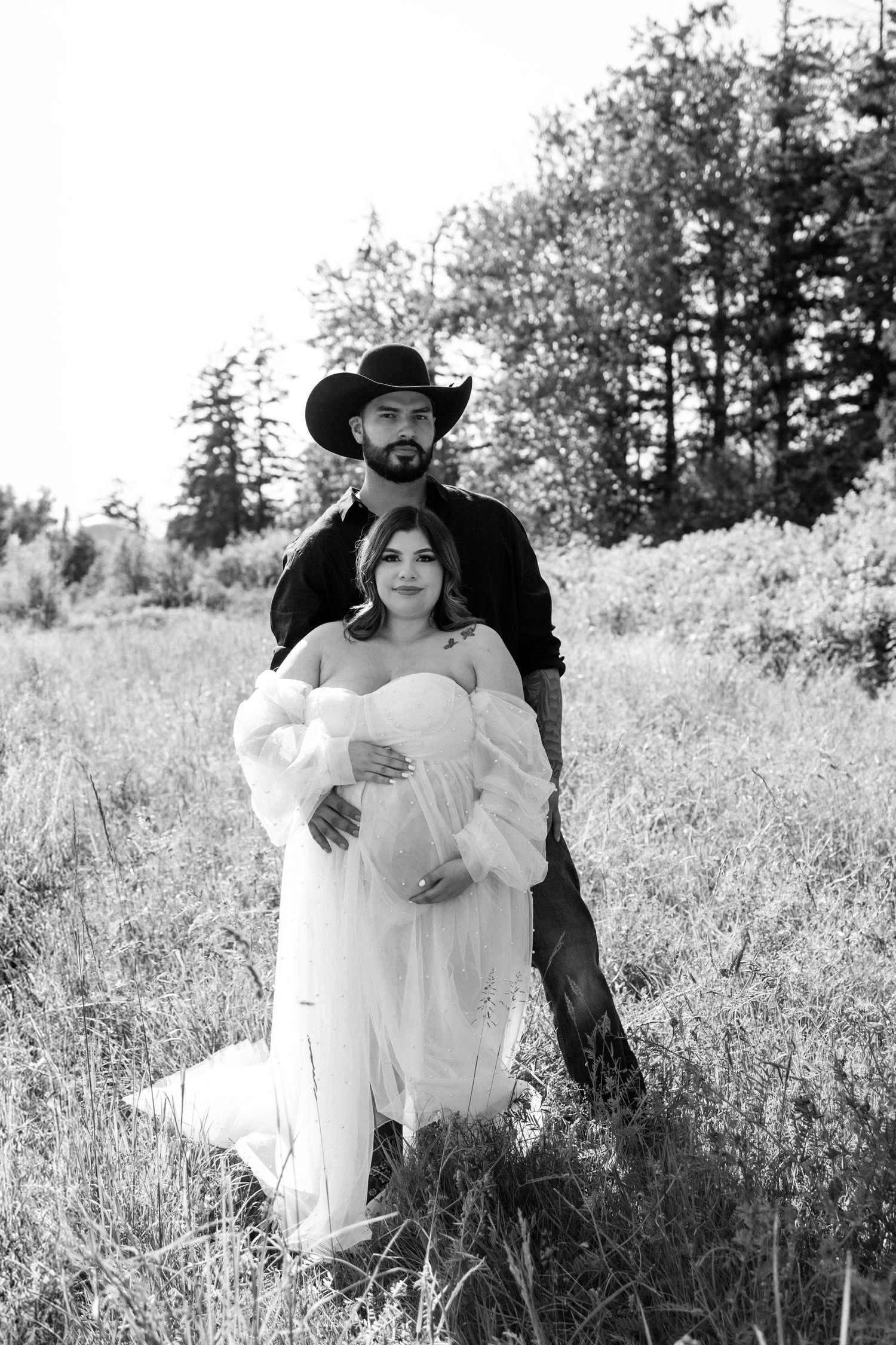 A couple poses in a field, with the woman wearing a white dress and man in cowboy hat and black attire.