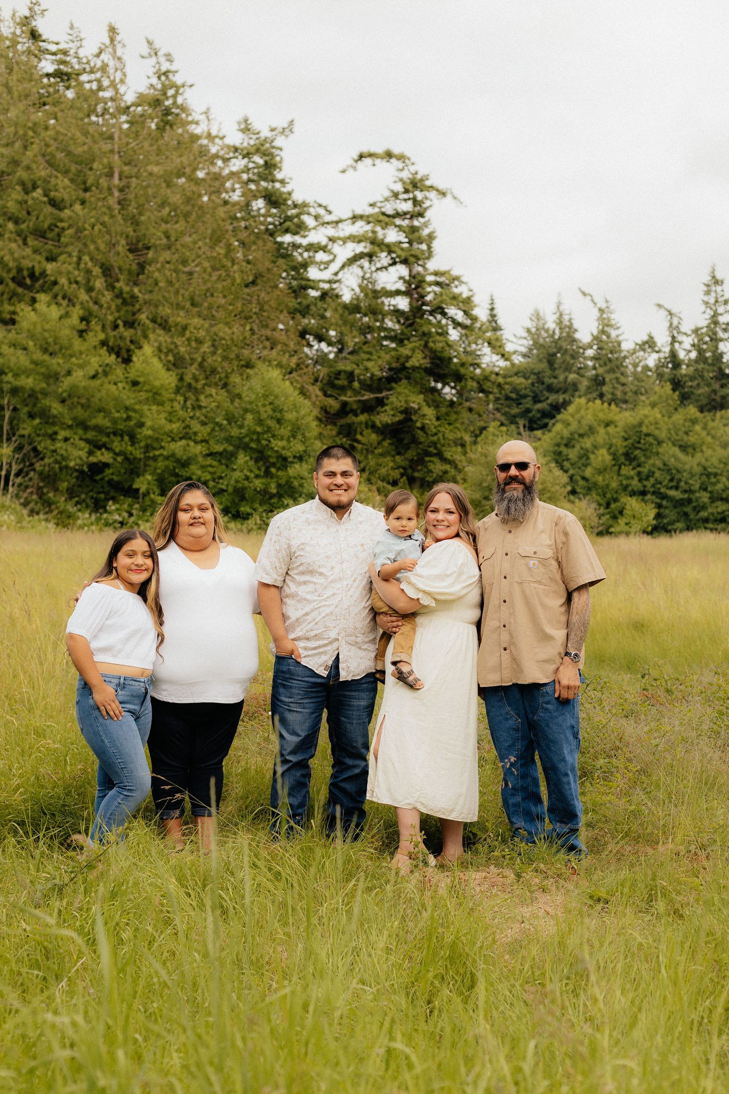 Family standing close together in a grassy field on a sunny summer day, arranged in playful poses.