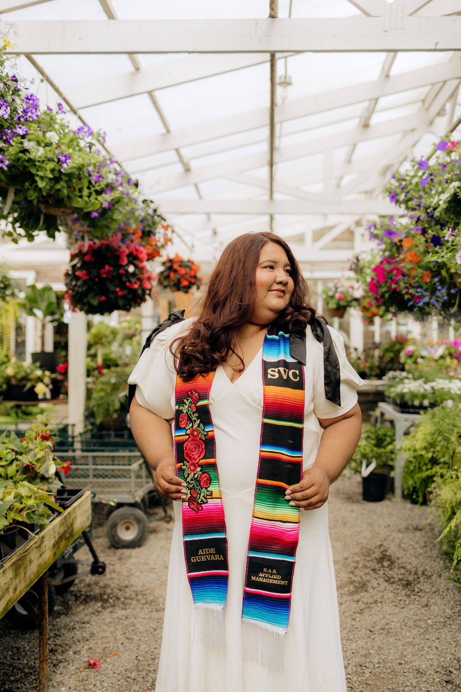 Shoppers browse colorful plants and hanging baskets inside a bright greenhouse with white beamed ceiling.