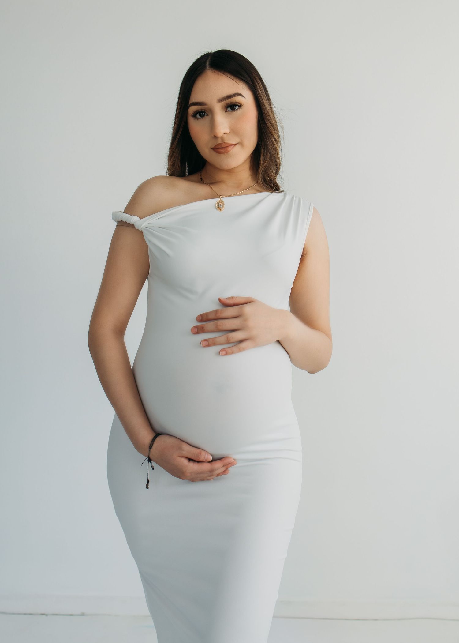 Woman in white form-fitting dress posing against plain wall during maternity photoshoot.