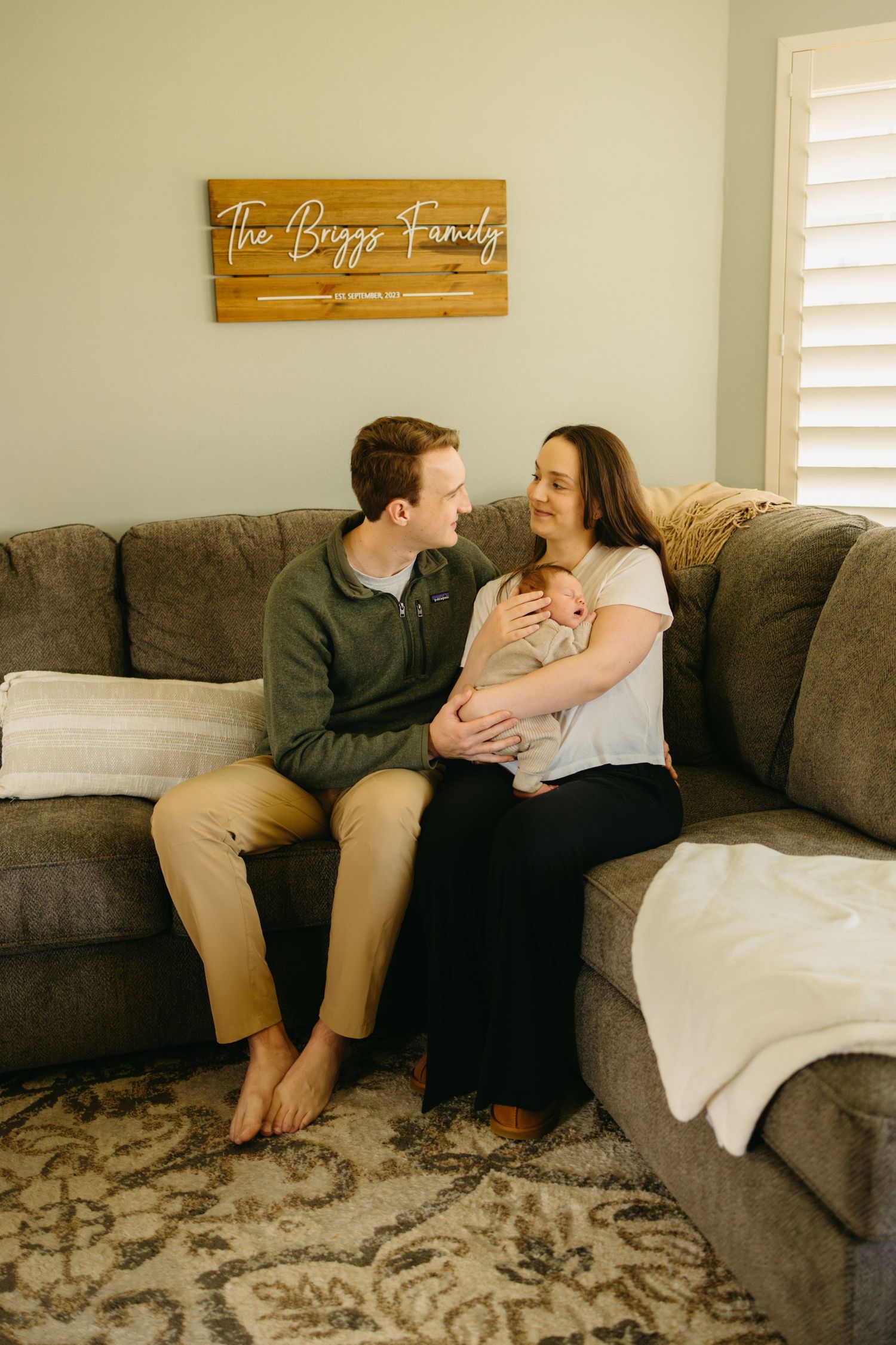 A couple sits together on a brown sectional sofa beneath a wooden sign, sharing a joyful moment at home.