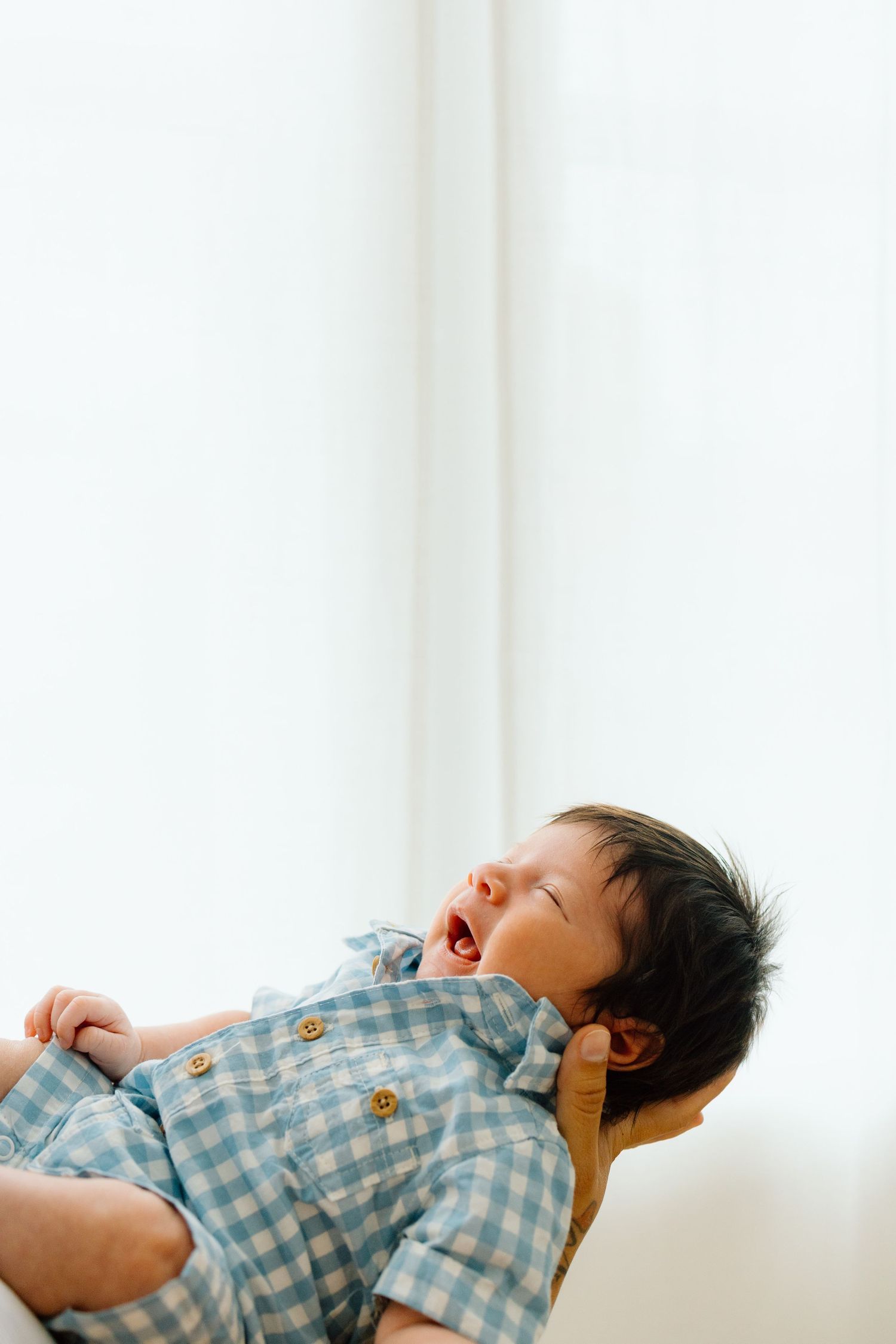 A laughing baby in a blue checkered shirt lays on a white background captured from above.
