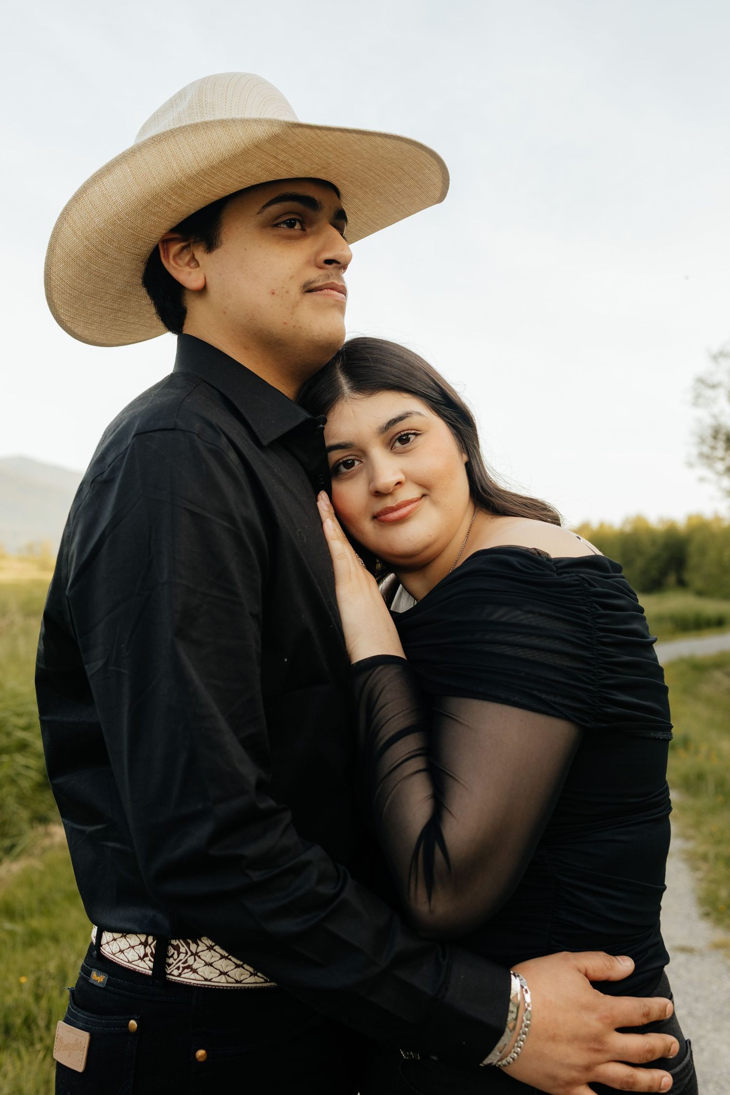 A couple embraces outdoors while wearing coordinated black outfits, with one wearing a western-style hat in a grassy setting.
