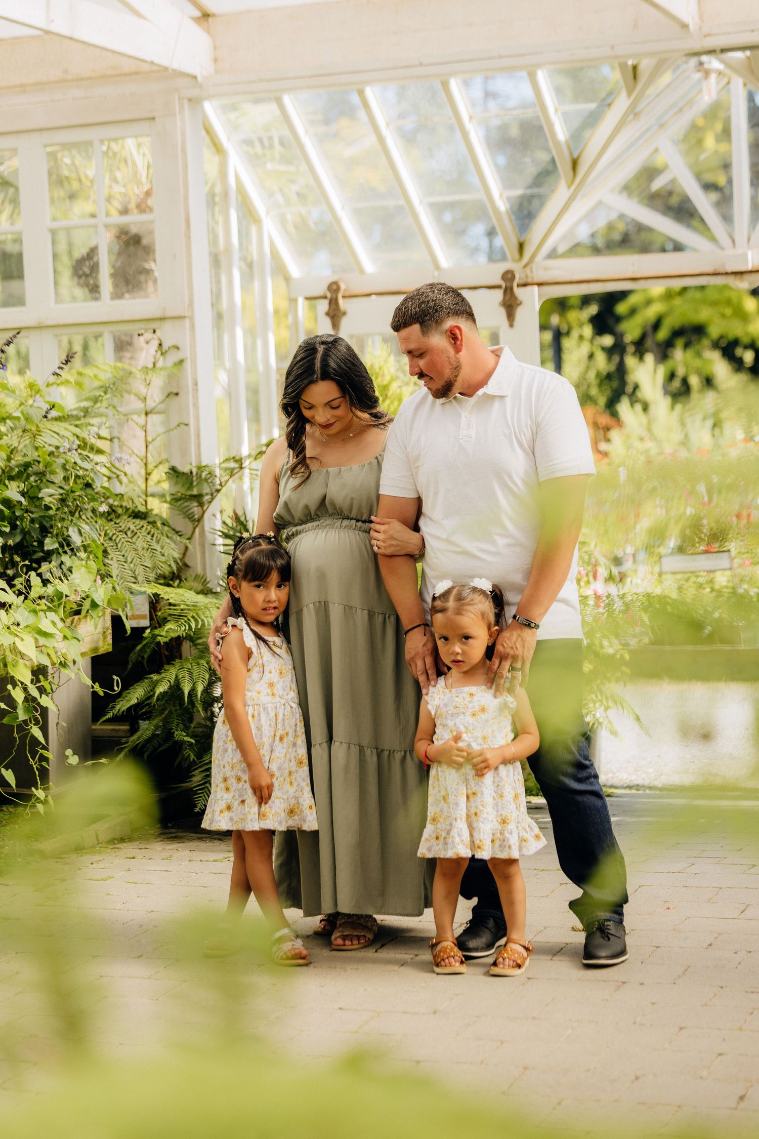 A family of four poses together in a bright greenhouse setting with white architectural details and lush greenery.