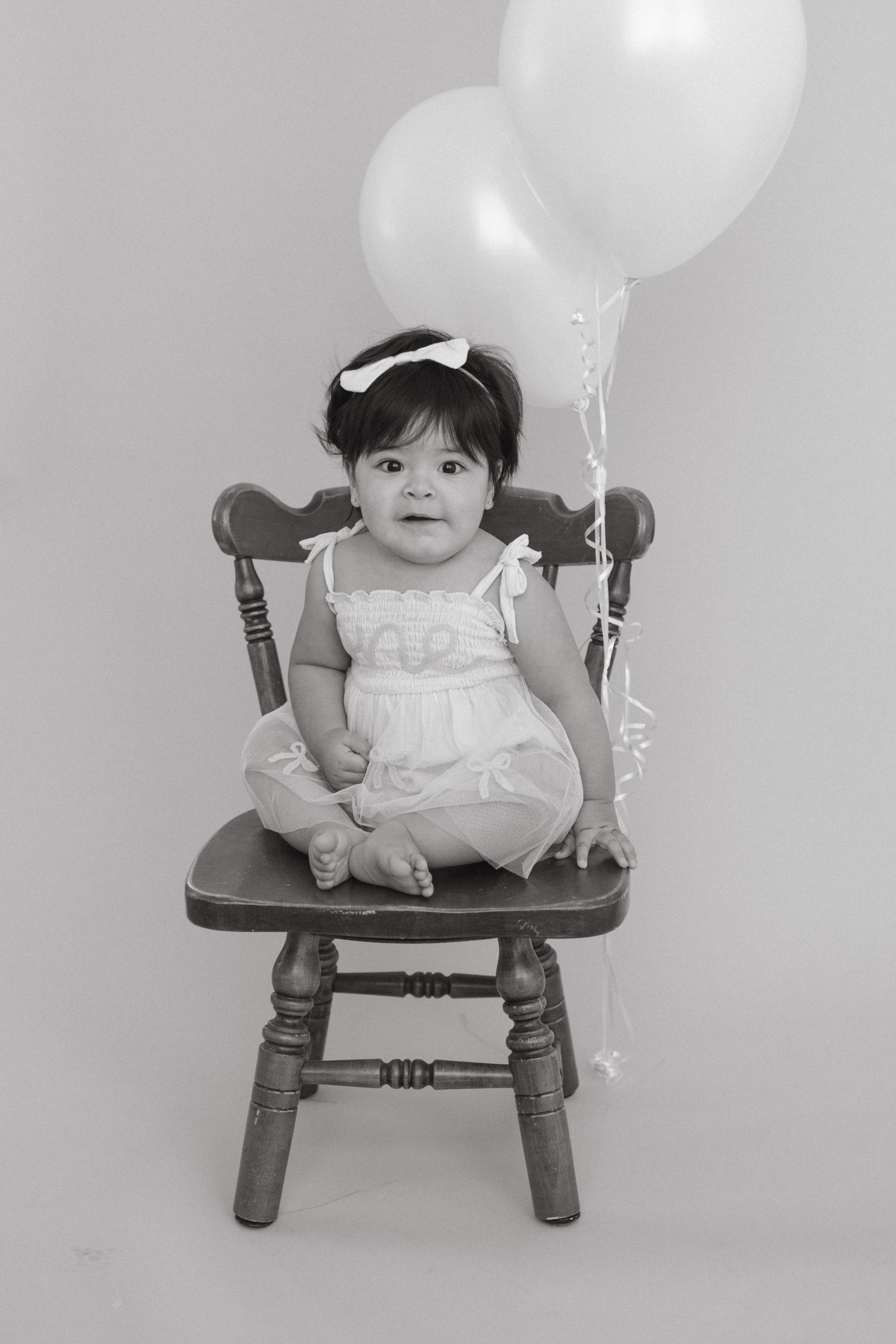Black and white portrait of a small child in a white dress seated on a wooden chair with balloons.