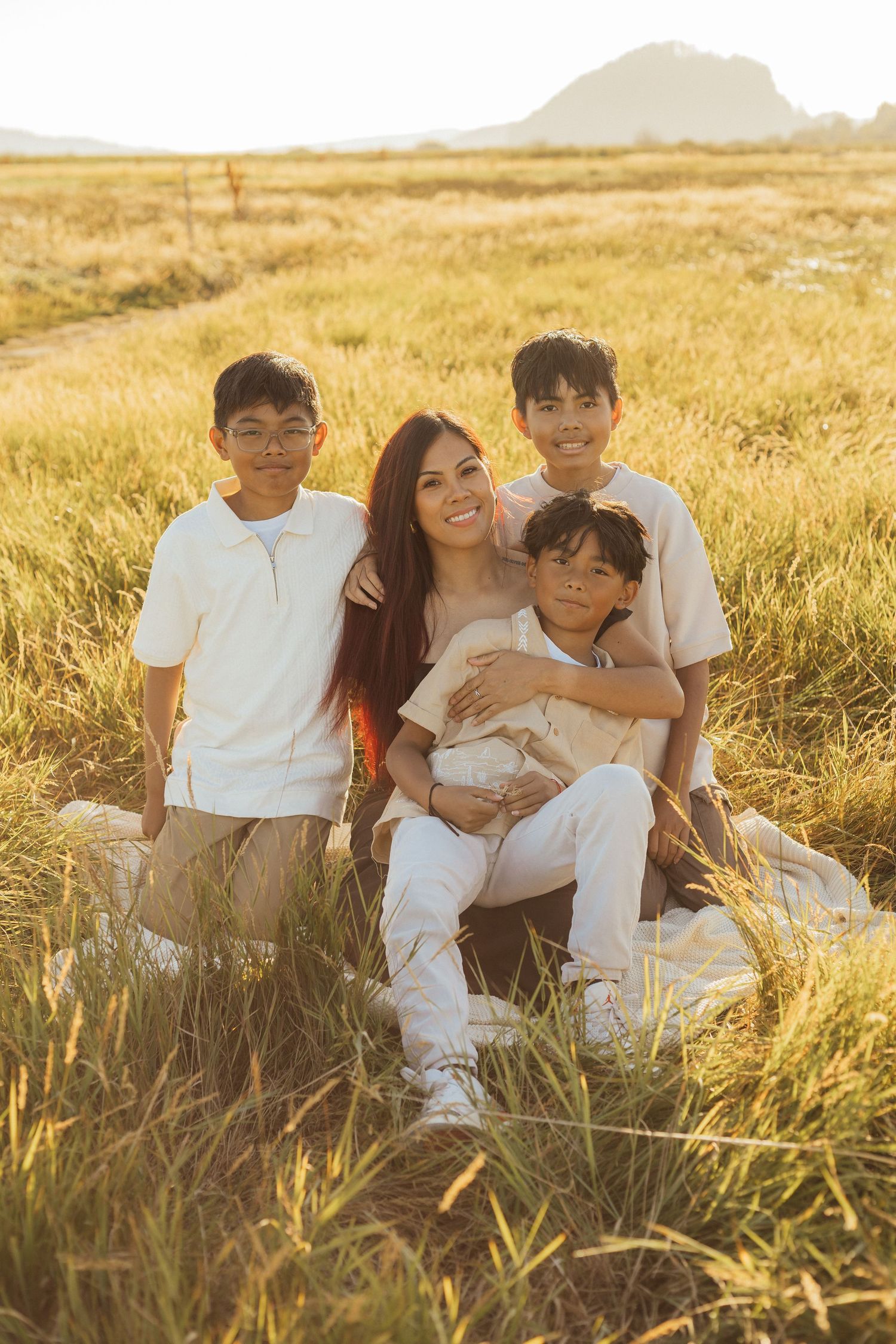 A family sits together in a golden wheat field at sunset with mountains visible in the background.