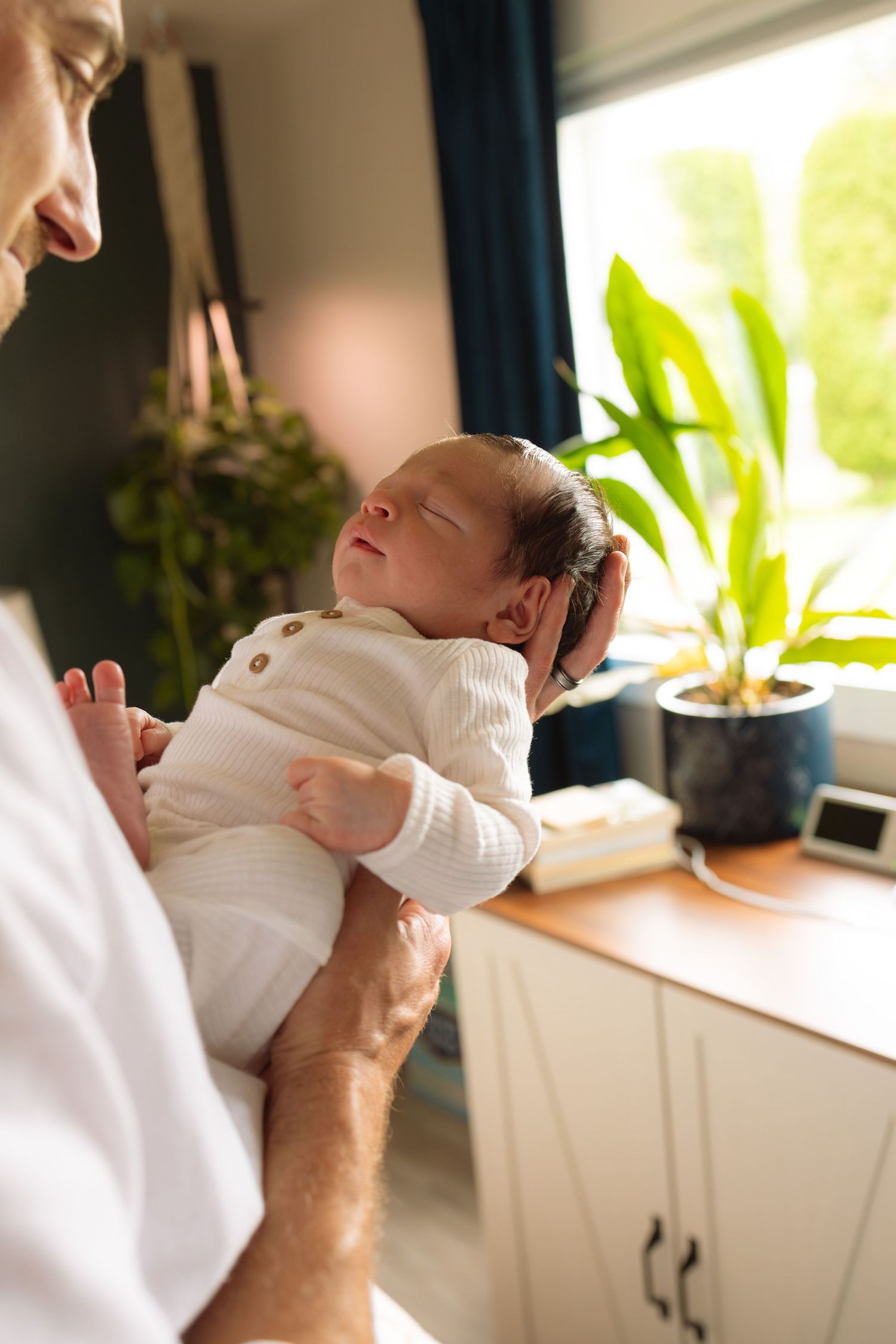 A sleeping newborn in a white sweater is cradled against someone's chest near a window with houseplants.