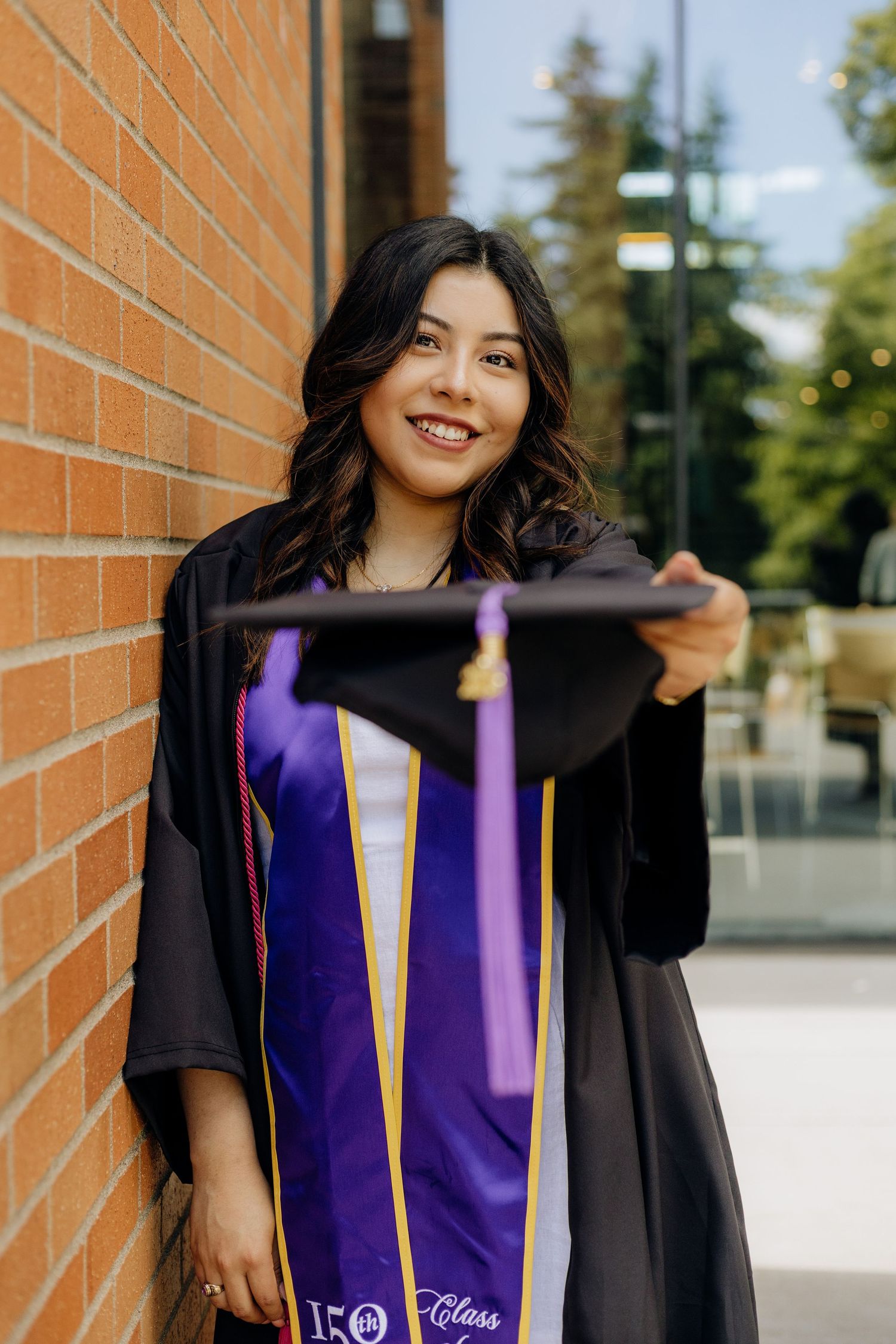 Graduate in black cap and gown with purple and gold sash smiles while posing against brick wall during commencement.