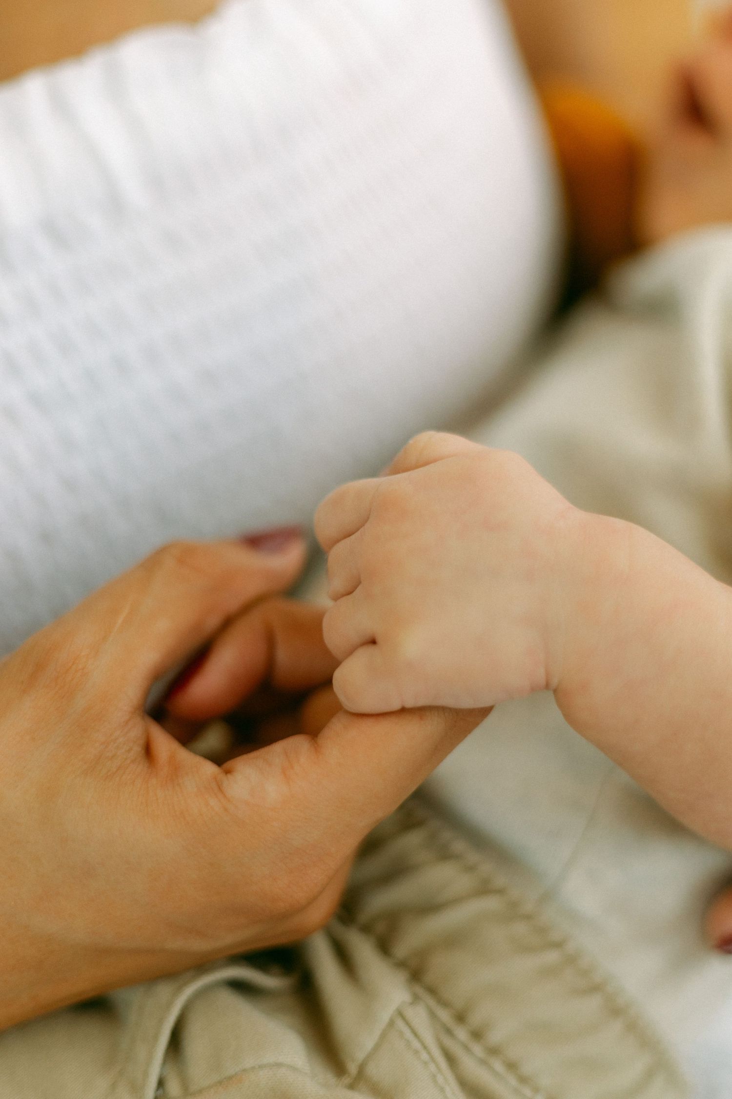 A close-up shot of an adult hand gently holding a tiny baby's hand against a soft beige background.