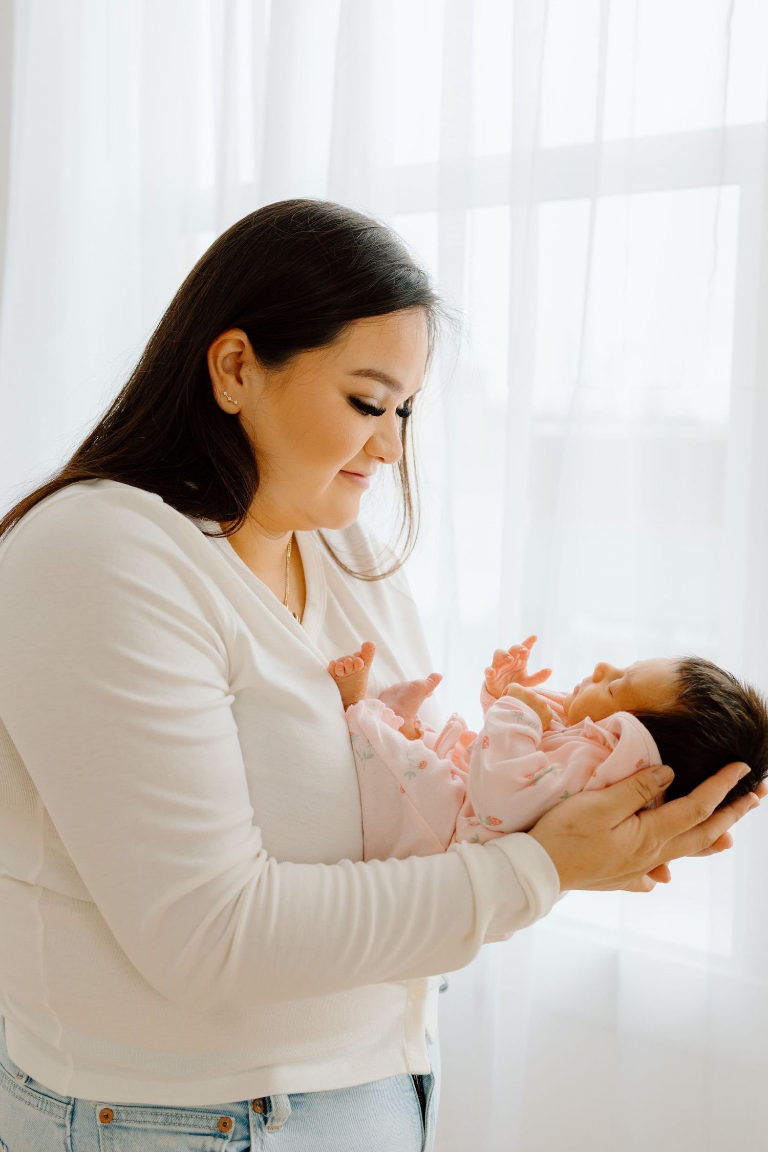 Mother in white sweater and jeans gently cradling newborn baby near bright window.