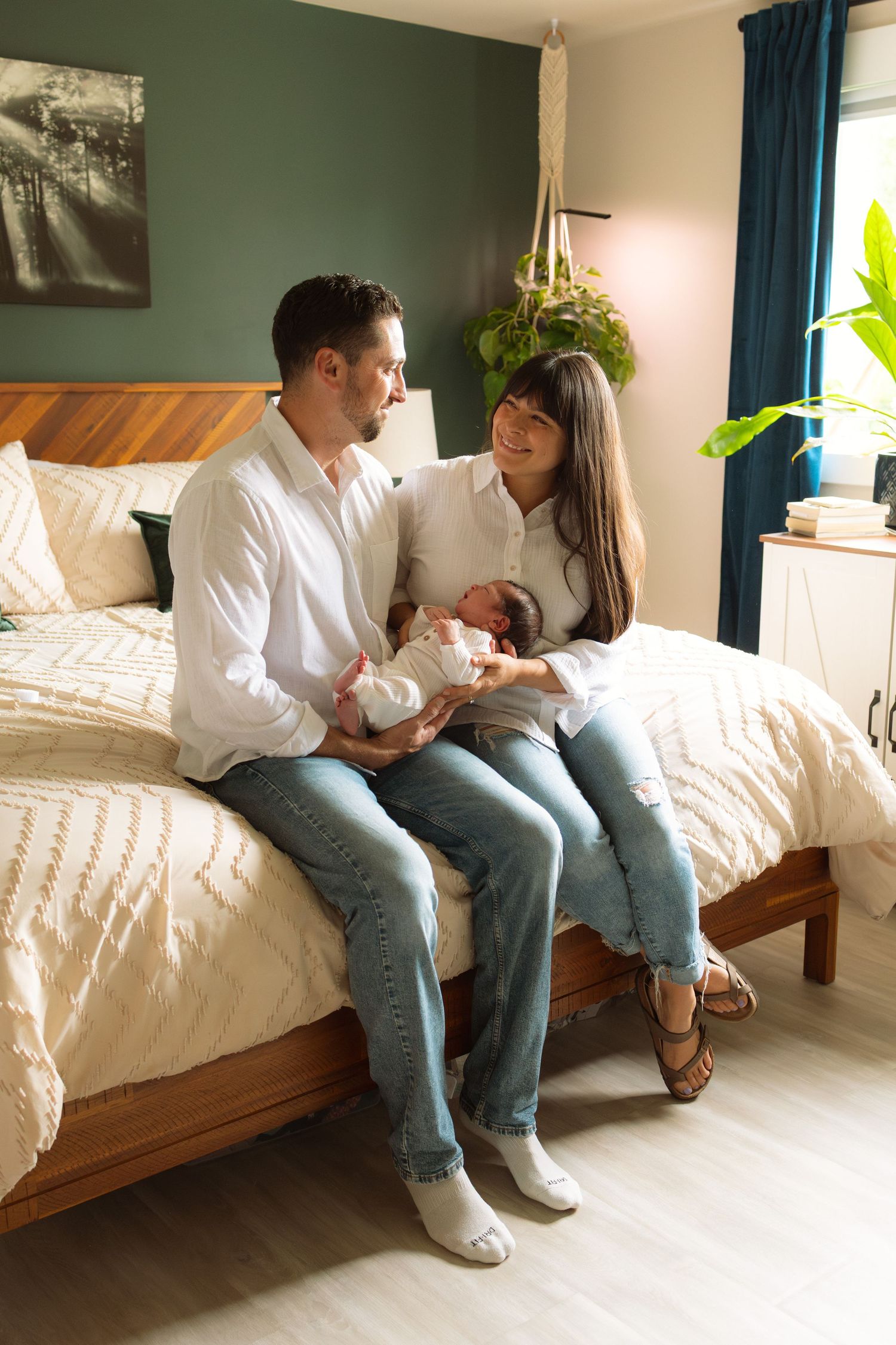 Happy couple sitting on bed holding newborn baby in cozy bedroom with green accent wall and teal curtains.