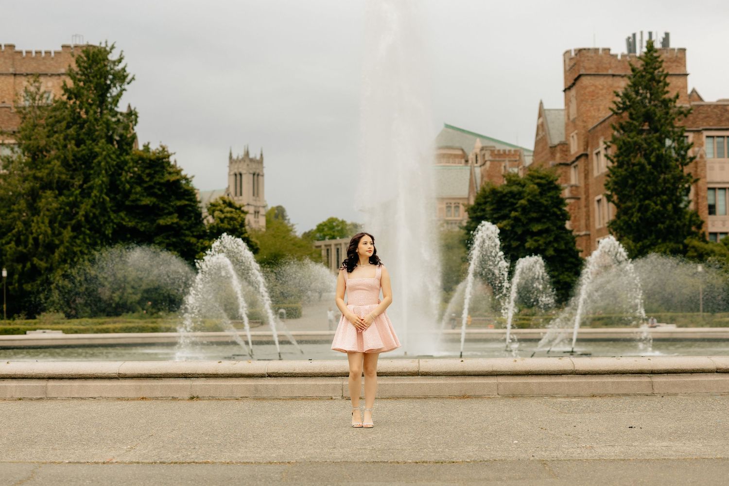 A figure stands in a pink dress in front of cascading water fountains on a university campus with historic buildings.
