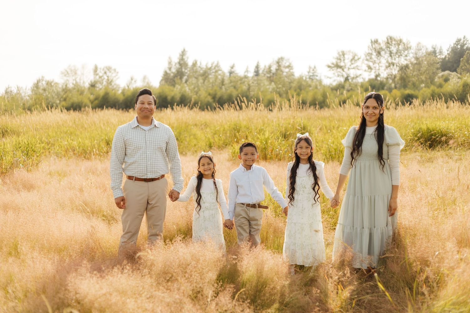 A family stands together in a golden sunlit meadow wearing coordinated light-colored outfits at sunset.