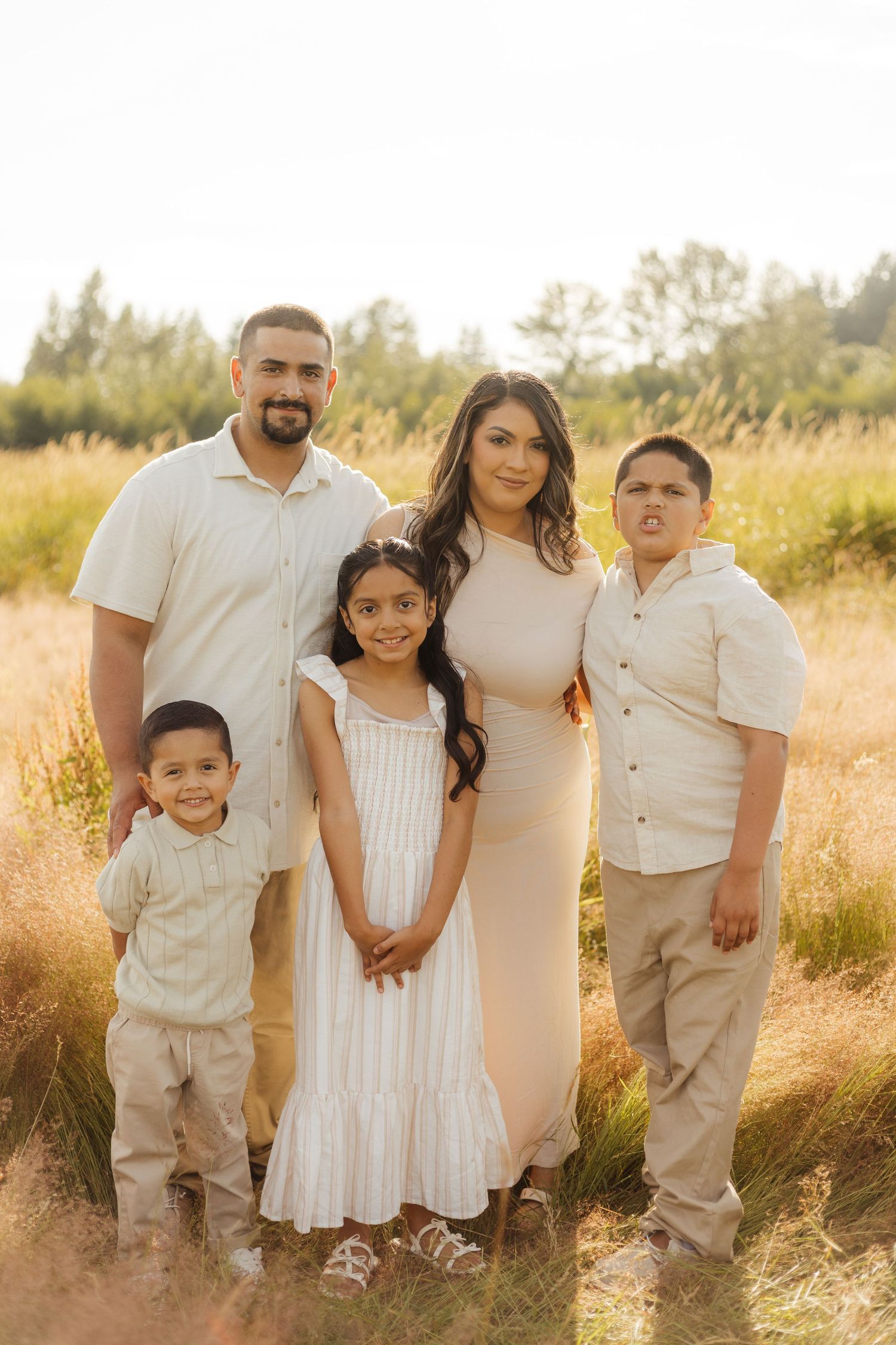 A family poses together outdoors wearing coordinated white and beige outfits in a golden grassy field at sunset.