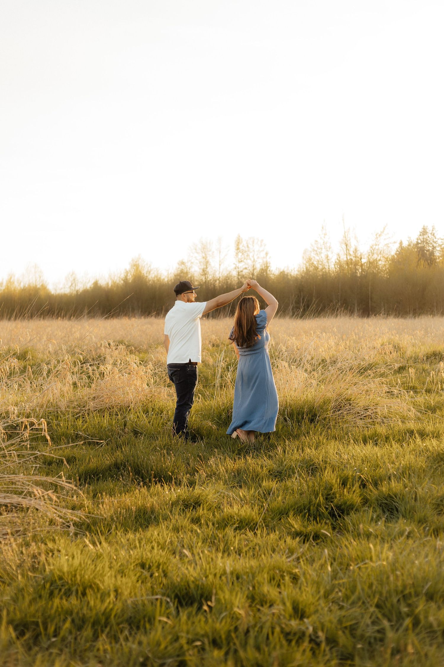 A couple dancing together in a golden sunlit meadow at sunset with tall grass and trees in the background.