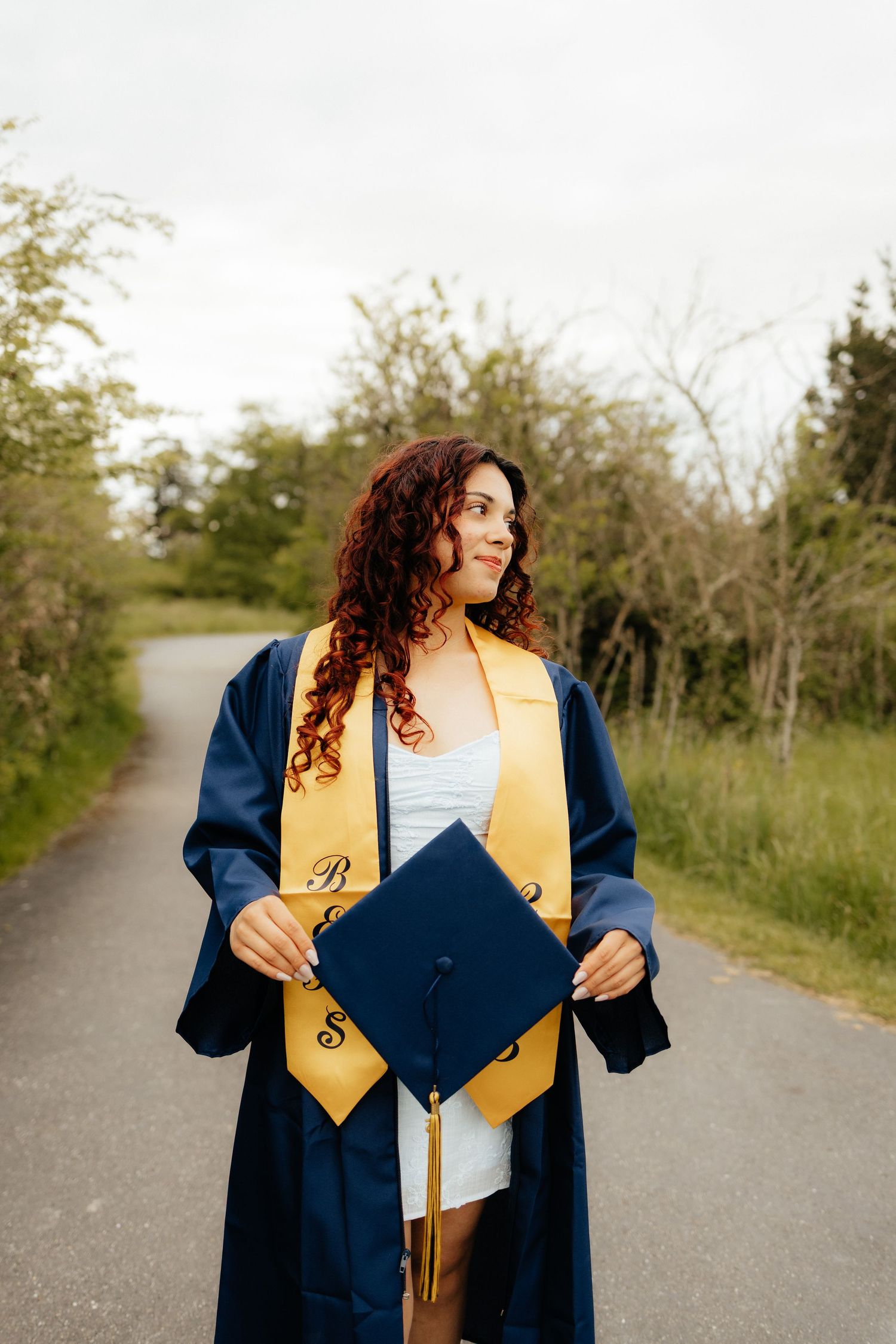 Graduate wearing navy blue and gold academic regalia holds diploma on outdoor pathway.