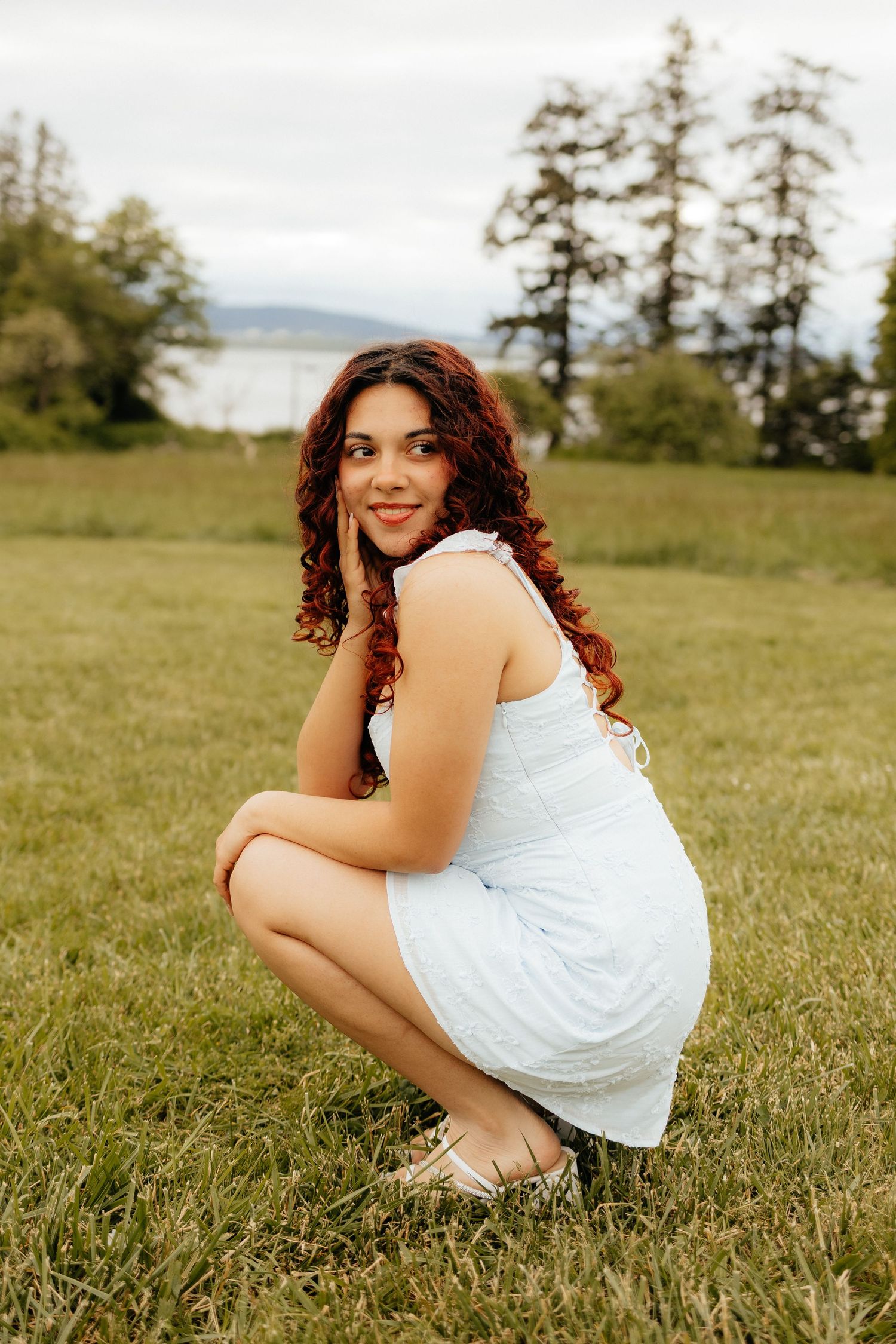 Person in white dress crouches in grassy meadow with evergreen trees and water view in background.