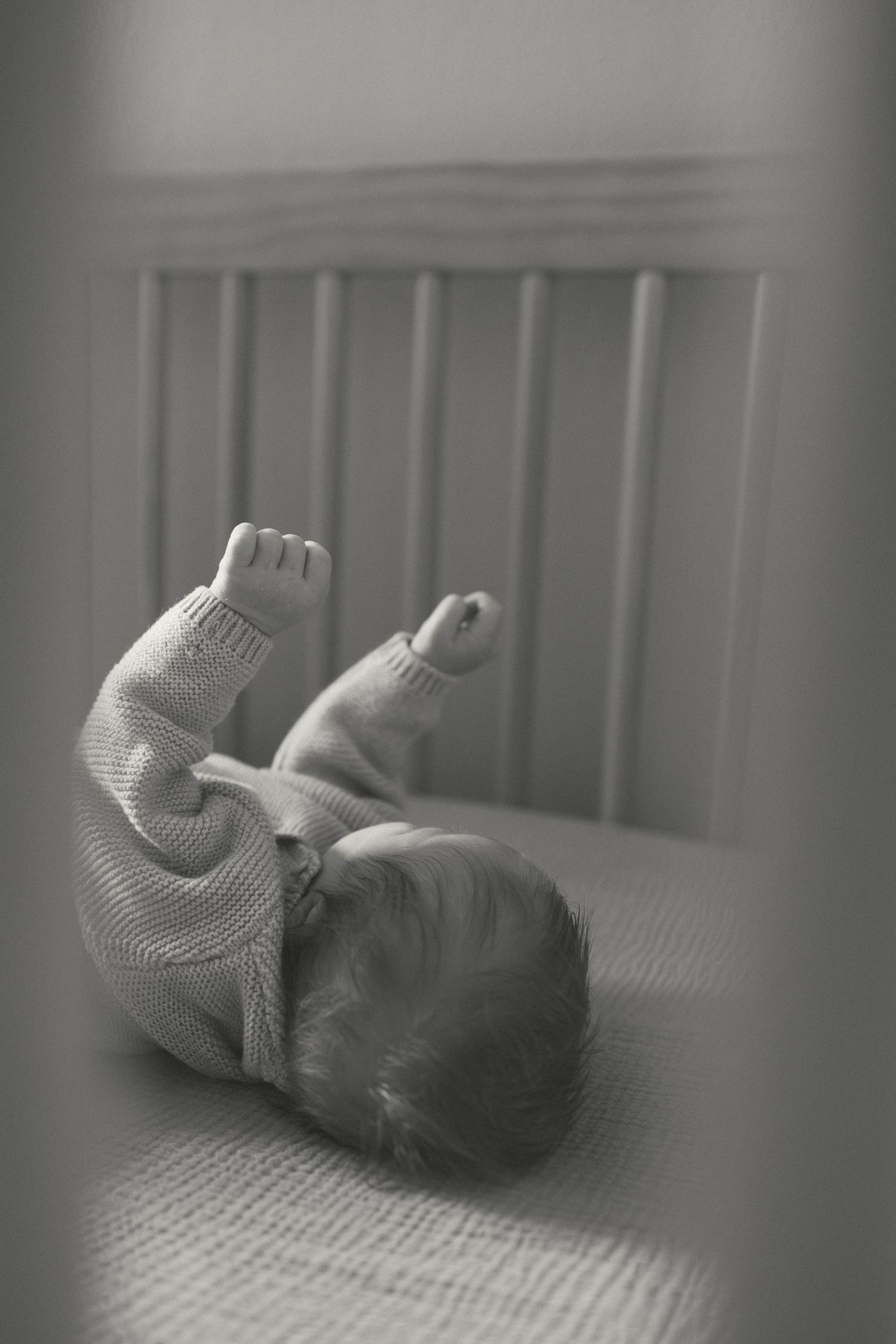 Black and white image of a sleeping baby wearing a knit sweater lying on a textured surface near crib bars.