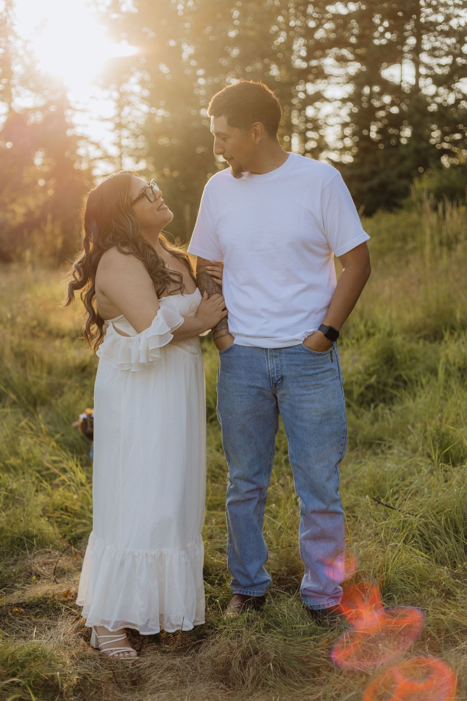 A romantic couple stands in a sun-dappled field wearing white dress and blue jeans during golden hour.