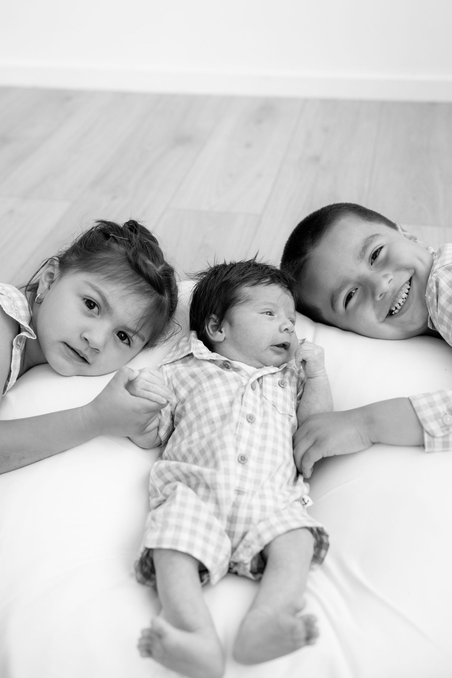 Three siblings lying together on white bedding in black and white portrait photograph.