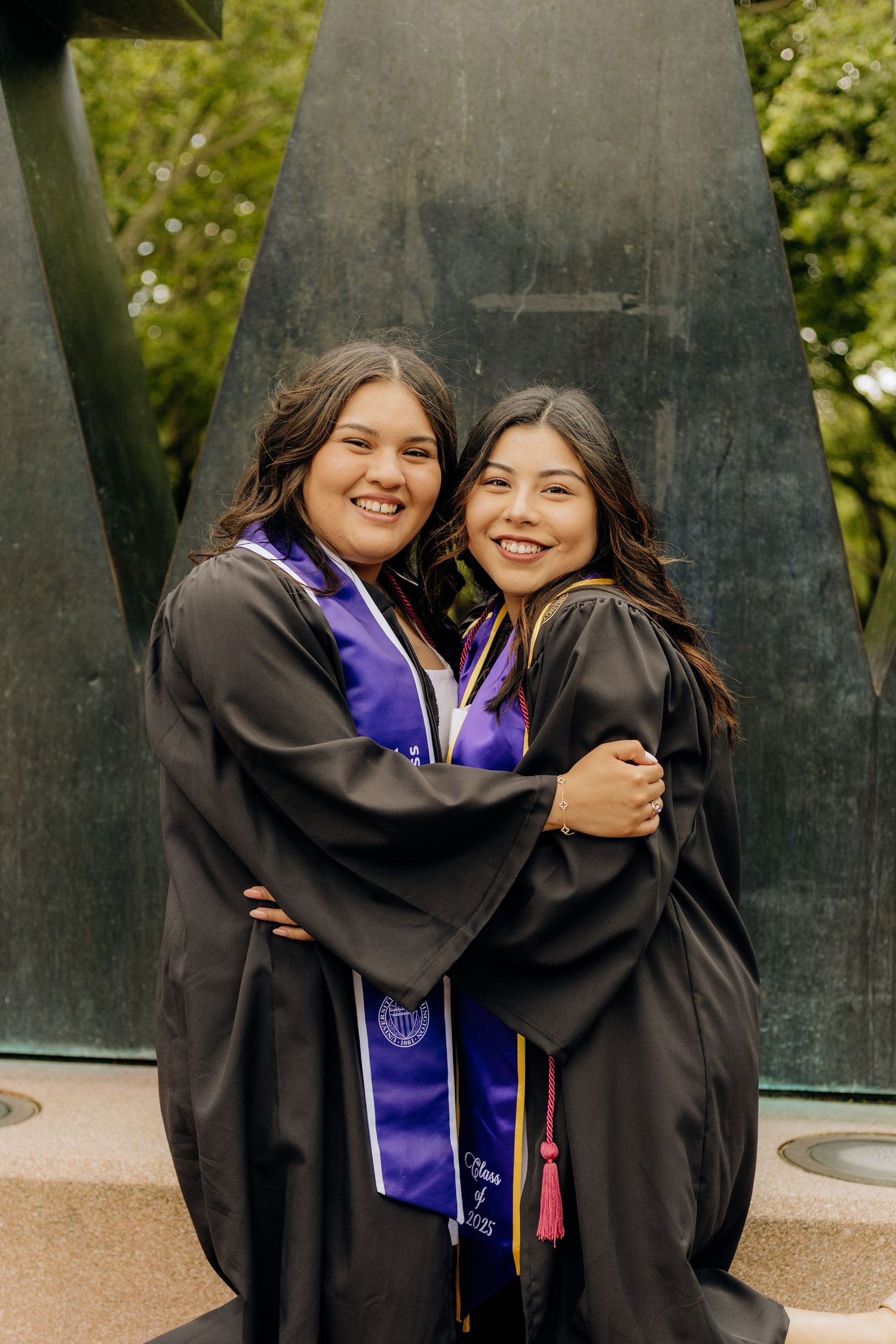 Two graduates embrace while wearing black graduation gowns and purple stoles in front of campus monument.