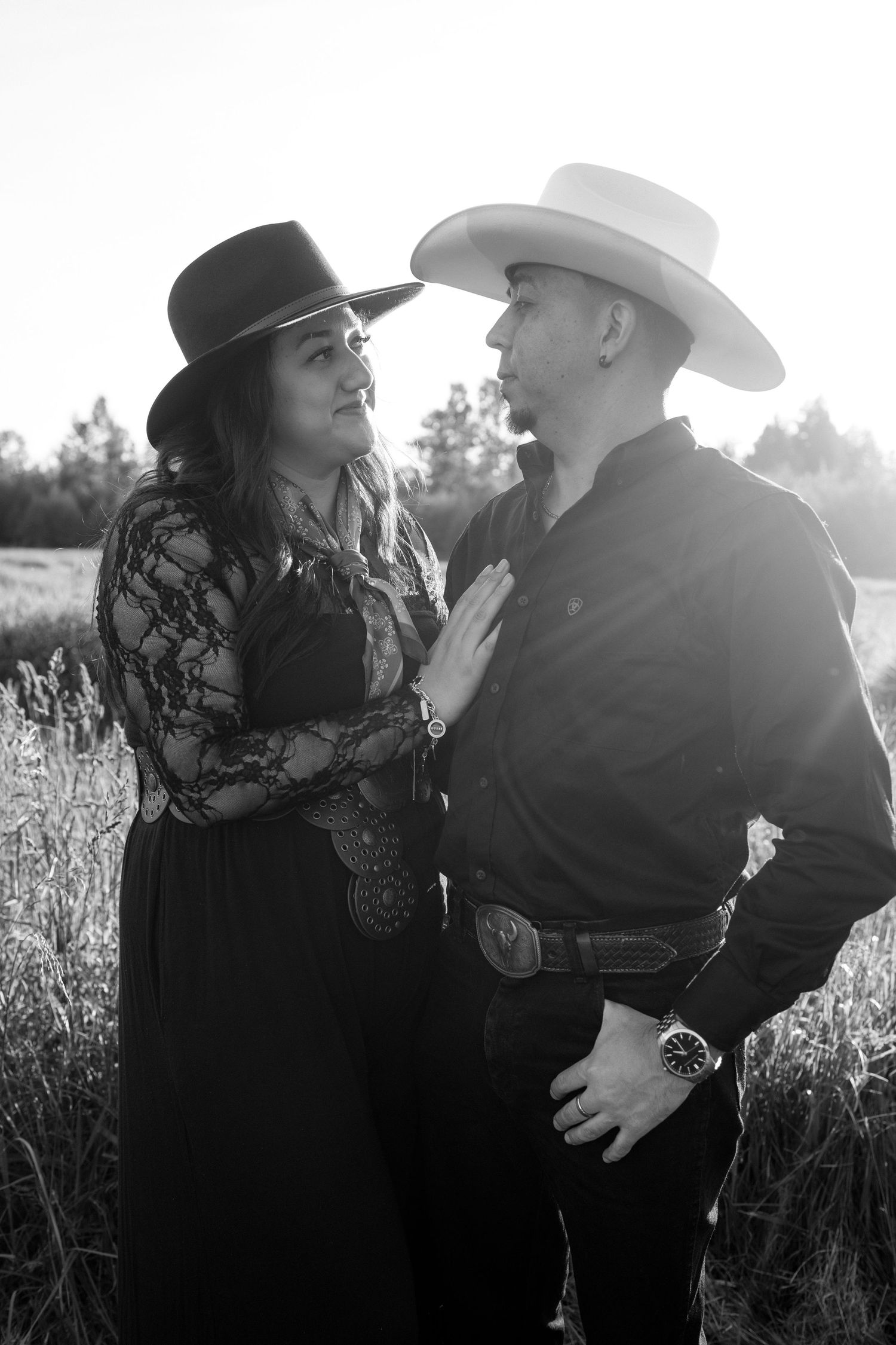 A romantic black and white portrait of a couple in western wear standing together in a sunlit field.