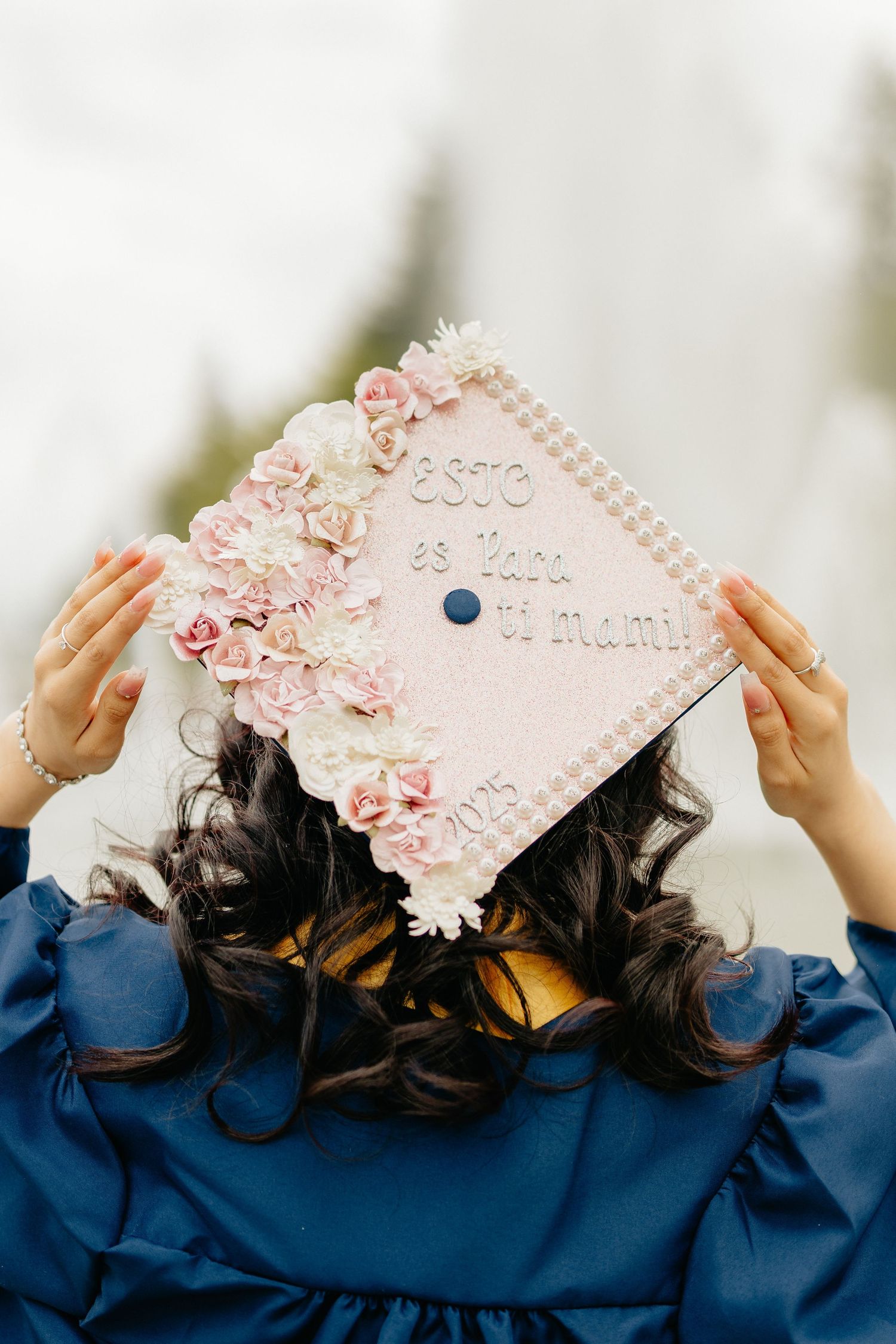 Graduate in navy blue gown holds up decorated graduation cap with pink flowers and pearl embellishments.