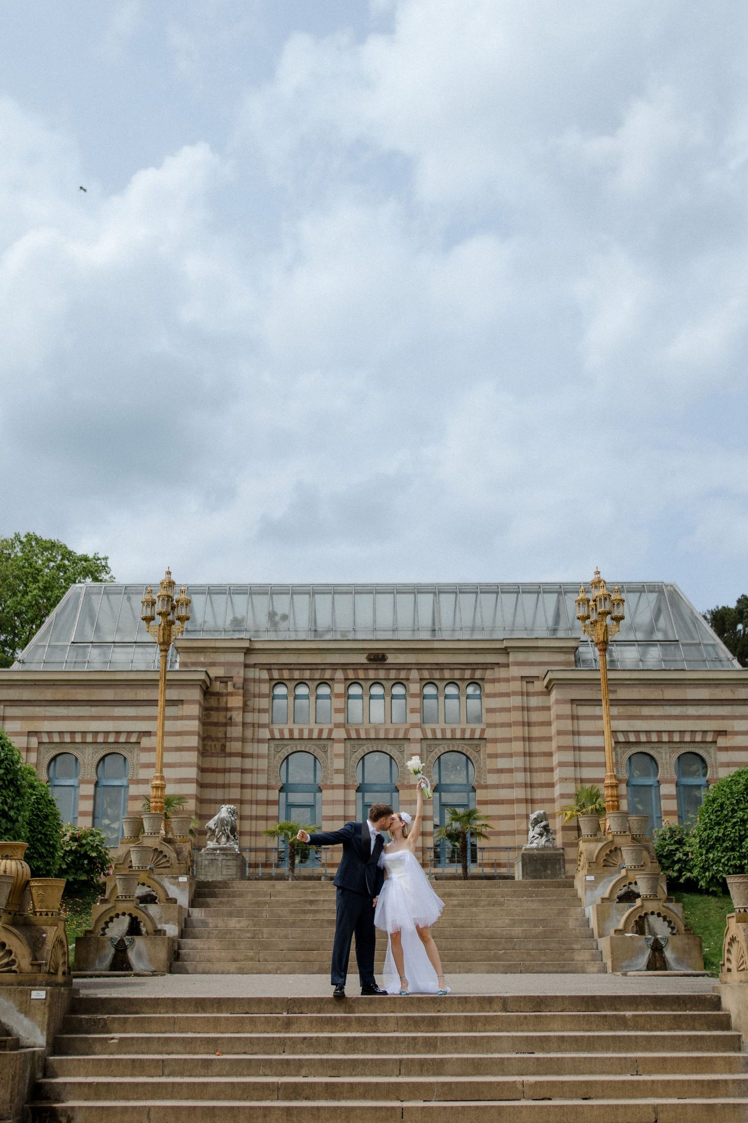 A couple stands on grand stone steps leading to a historic building with ornate architecture.