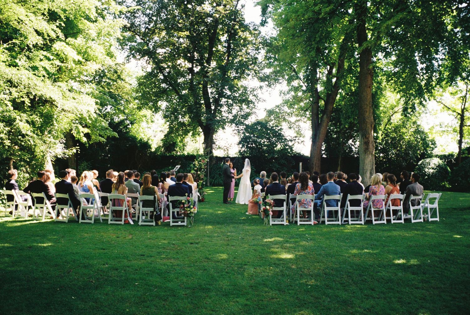 White chairs arranged in rows on a lush green lawn for an outdoor ceremony under towering trees.