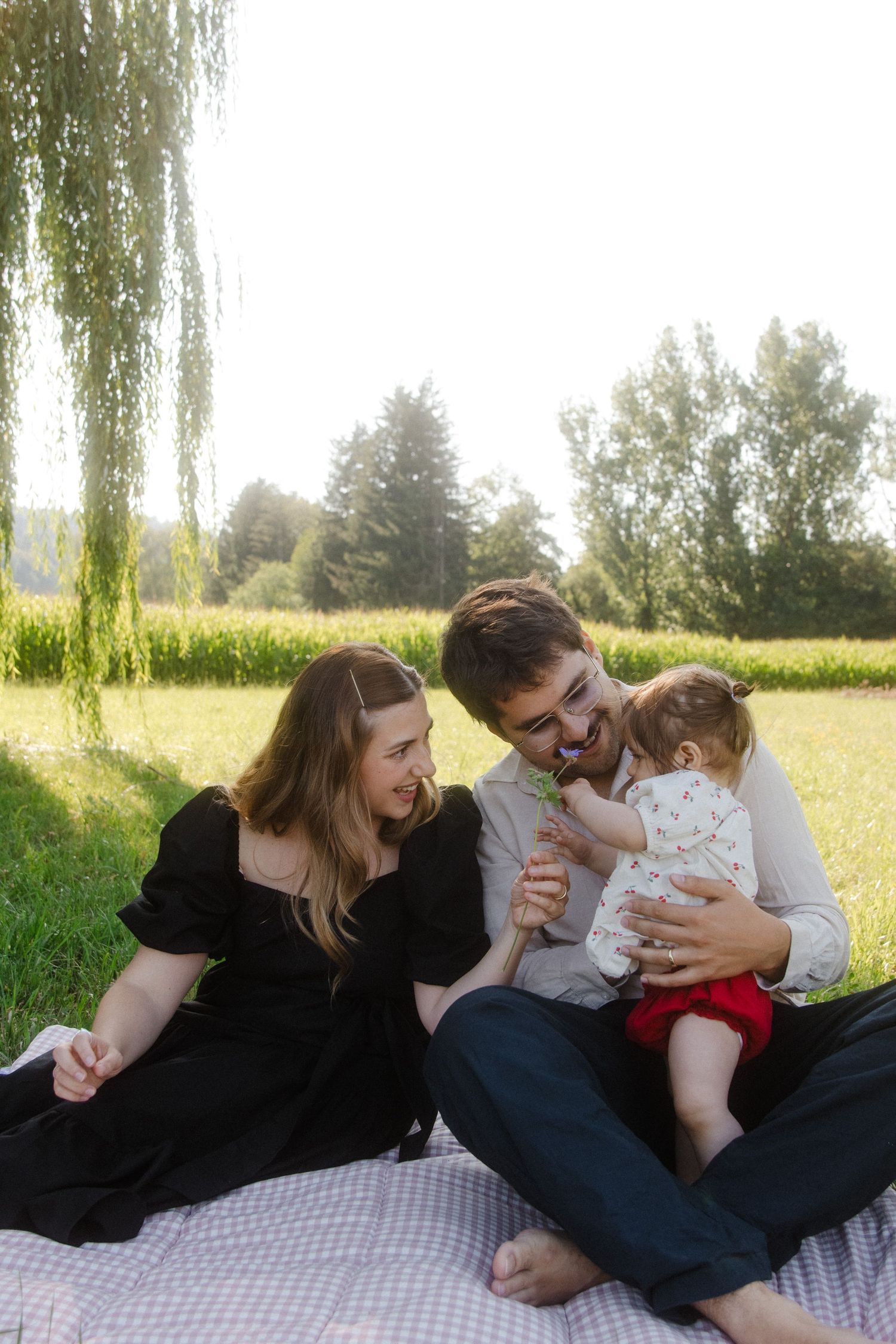 Young family enjoying a sunny park picnic on a blanket, parents smiling at baby being fed a treat outdoors.