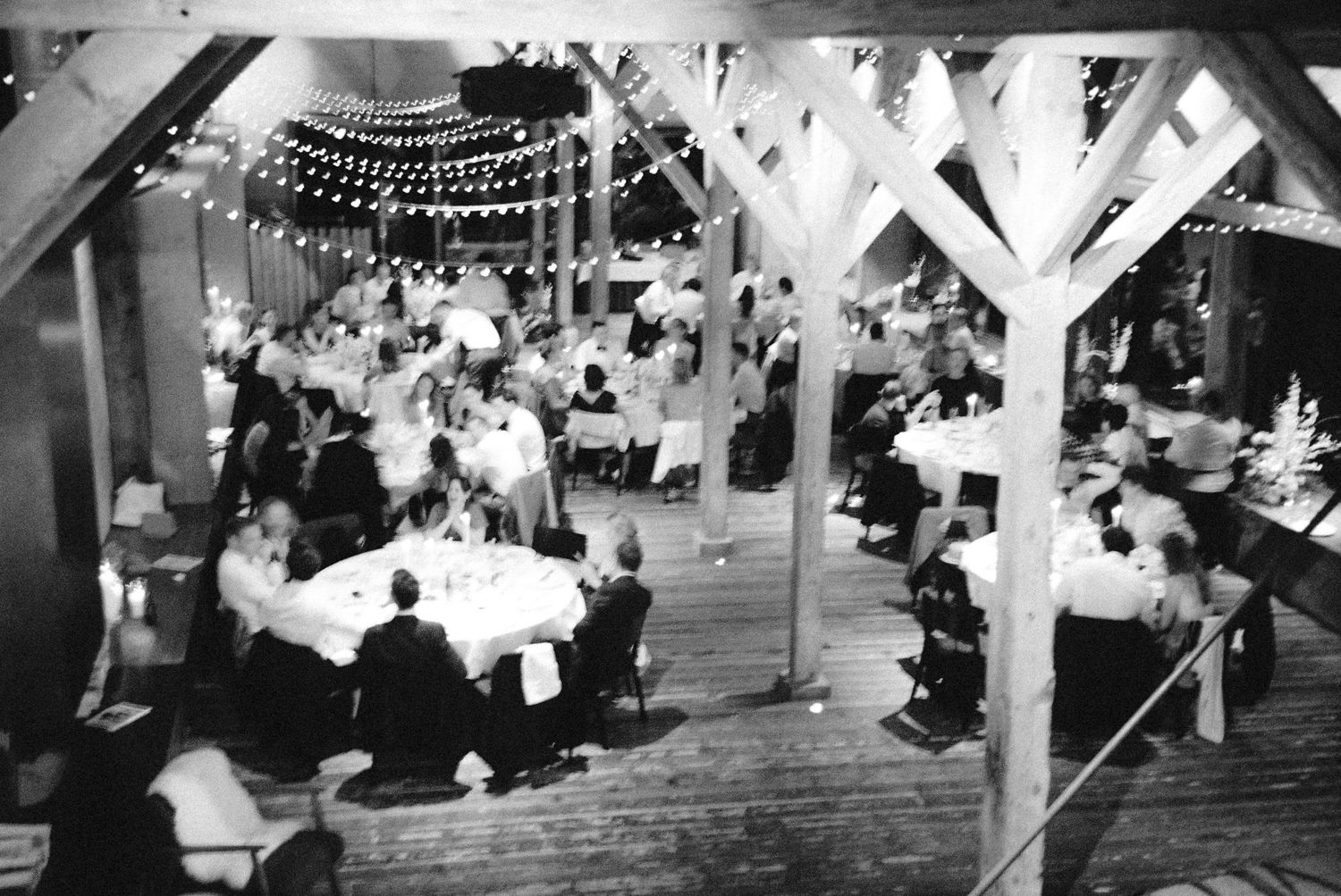 Black and white photo of elegant barn wedding reception with string lights, bunting, and guests seated at round tables.