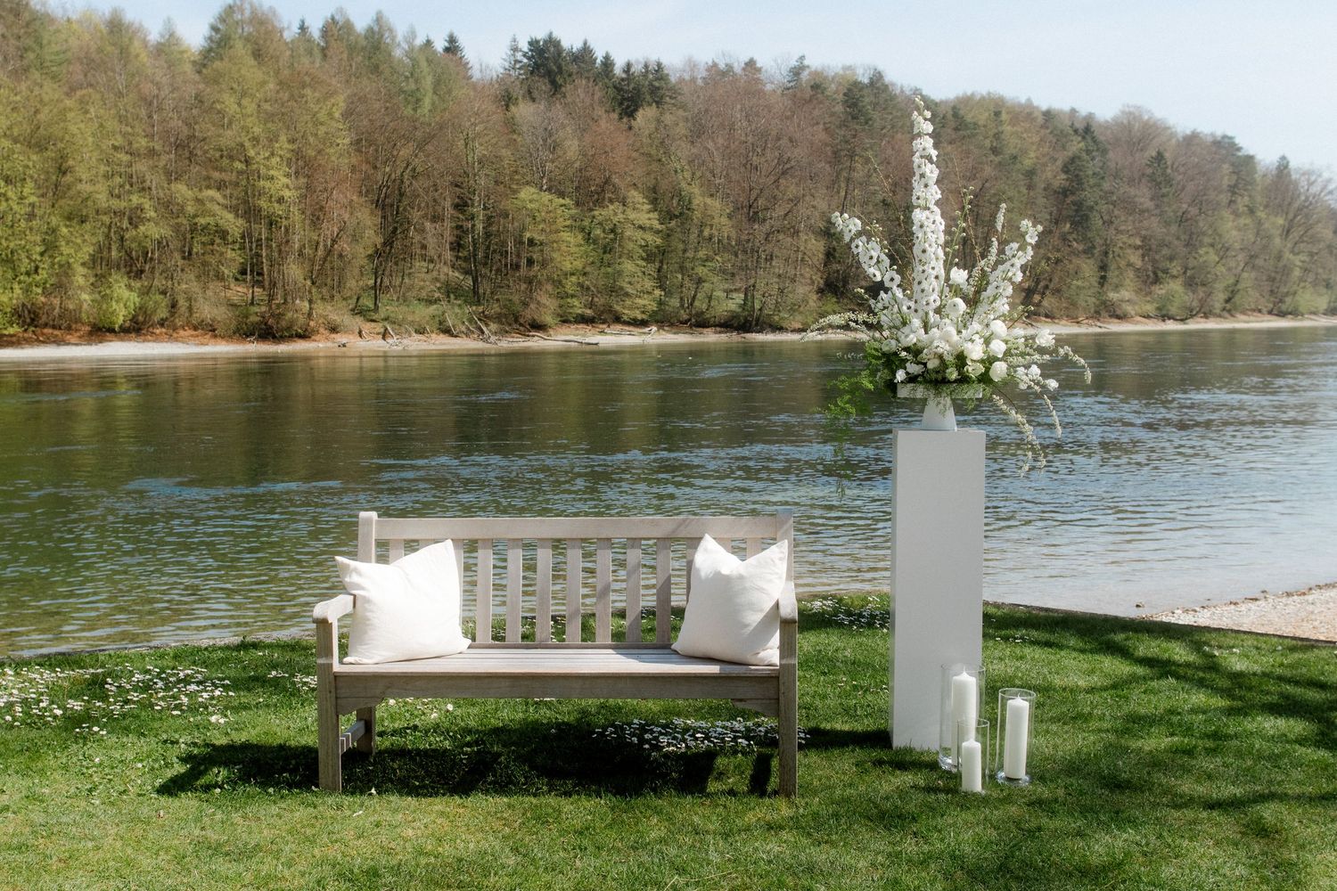 White bench and floral arrangement beside a serene river with lush green trees in background on a sunny day.