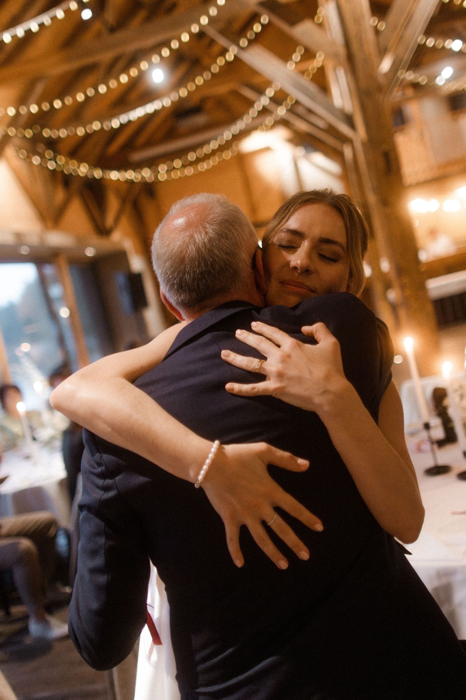 A woman in a navy dress embraces an older man in a suit during a wedding reception in a warmly lit barn.