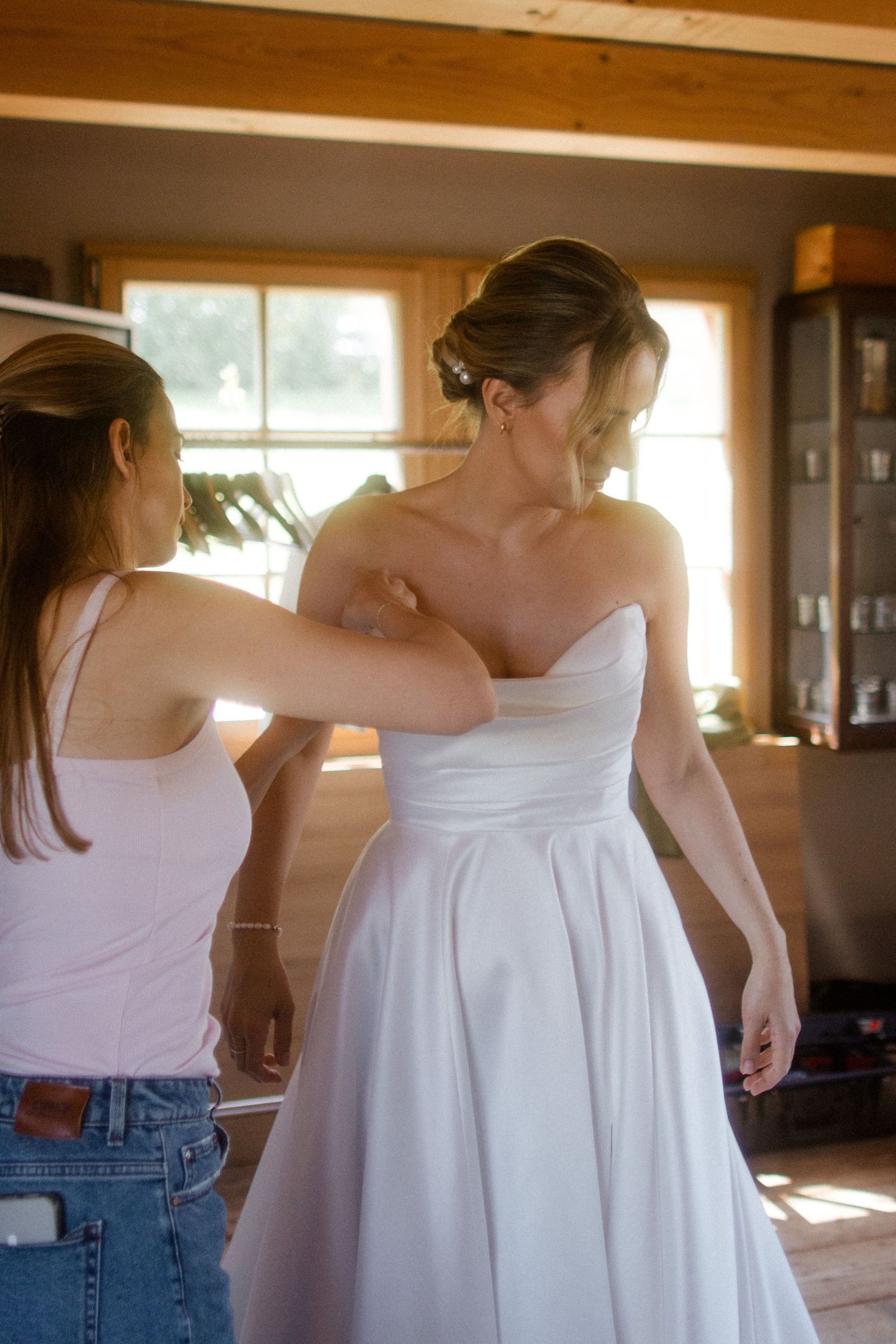Bridesmaid helping bride into elegant strapless white wedding gown in warmly lit rustic interior.