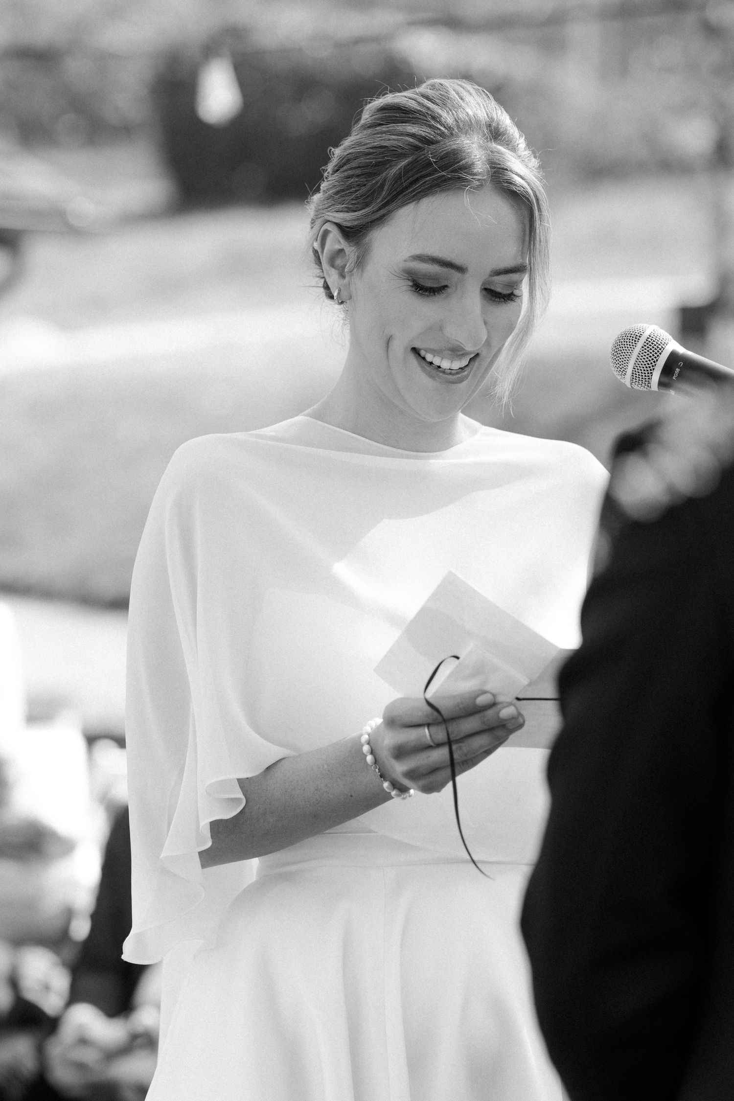 Black and white portrait of a smiling bride in white cape dress reading vows at outdoor wedding ceremony.
