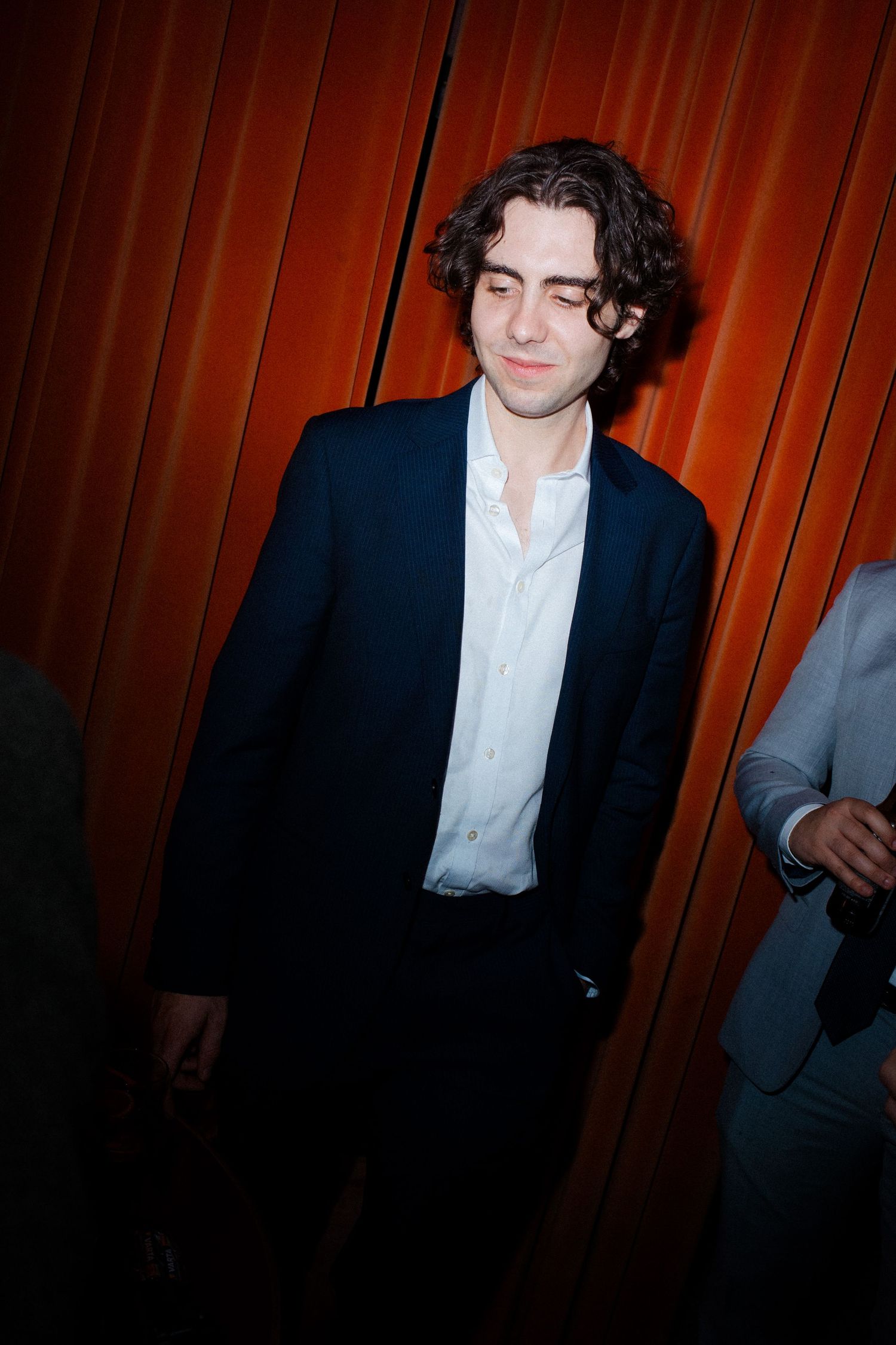 Young man in navy blazer and white shirt stands before an orange curtain at a formal event.