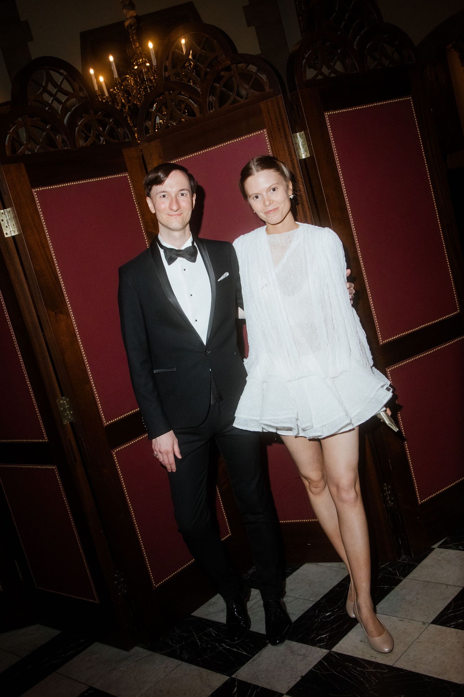 Elegant couple posing at formal event, man in black tuxedo, woman in white ruffled mini dress, ornate interior backdrop.