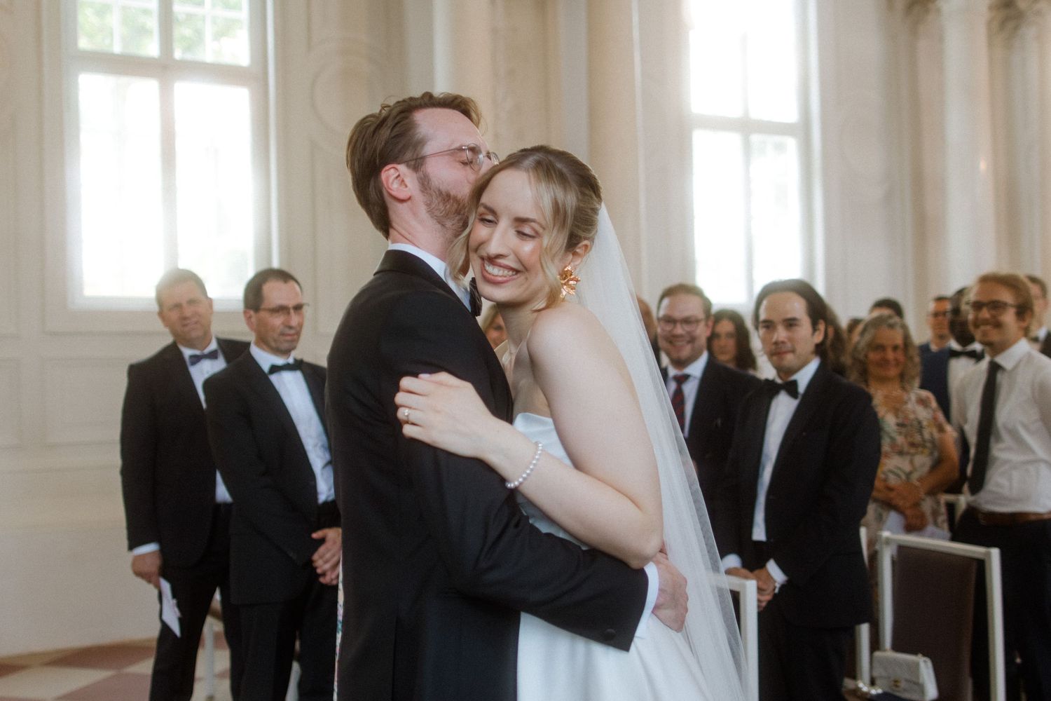 Intimate wedding moment in an ornate white hall with classical architecture.