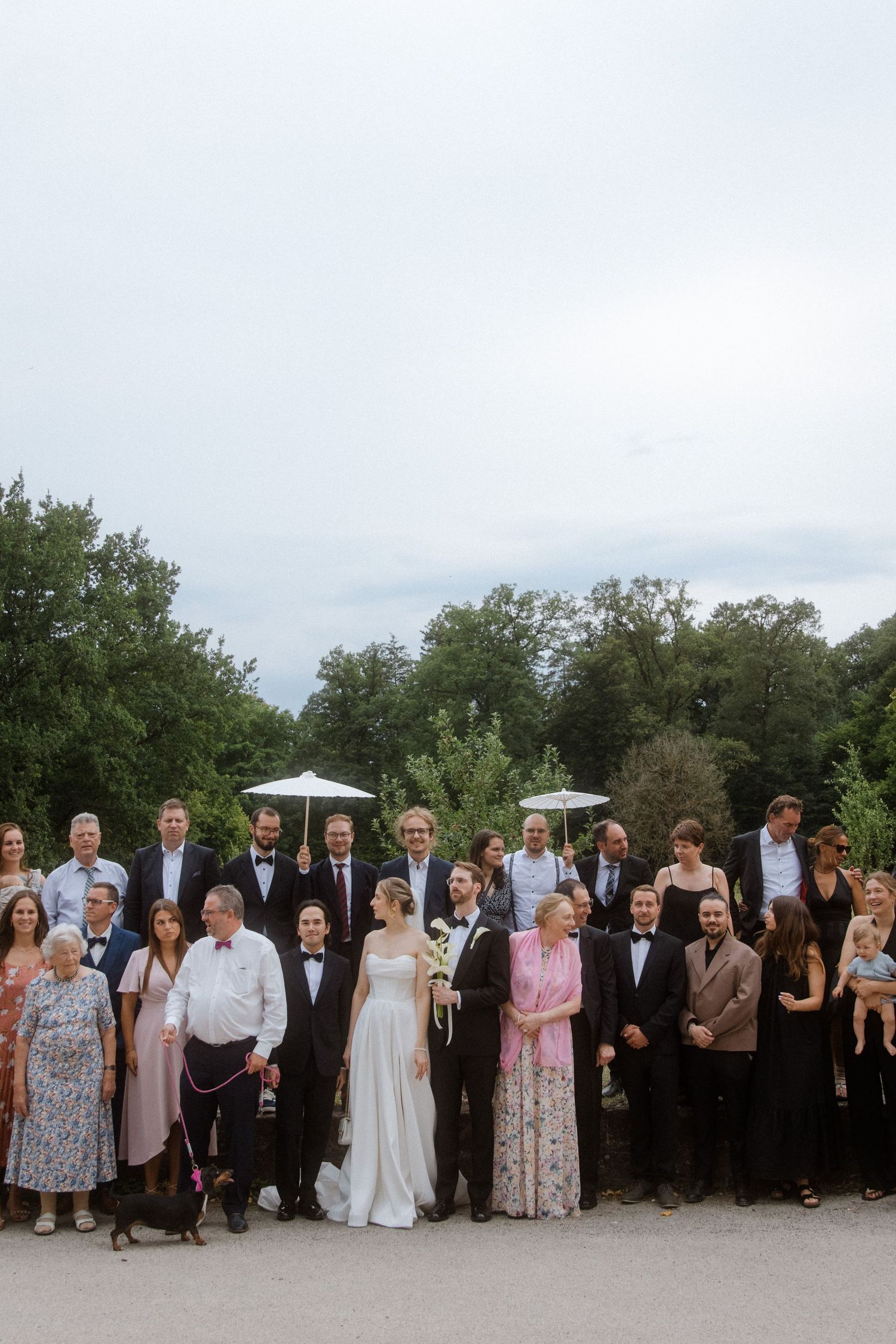 Wedding party and guests gather outdoors for formal group photos on a cloudy day.
