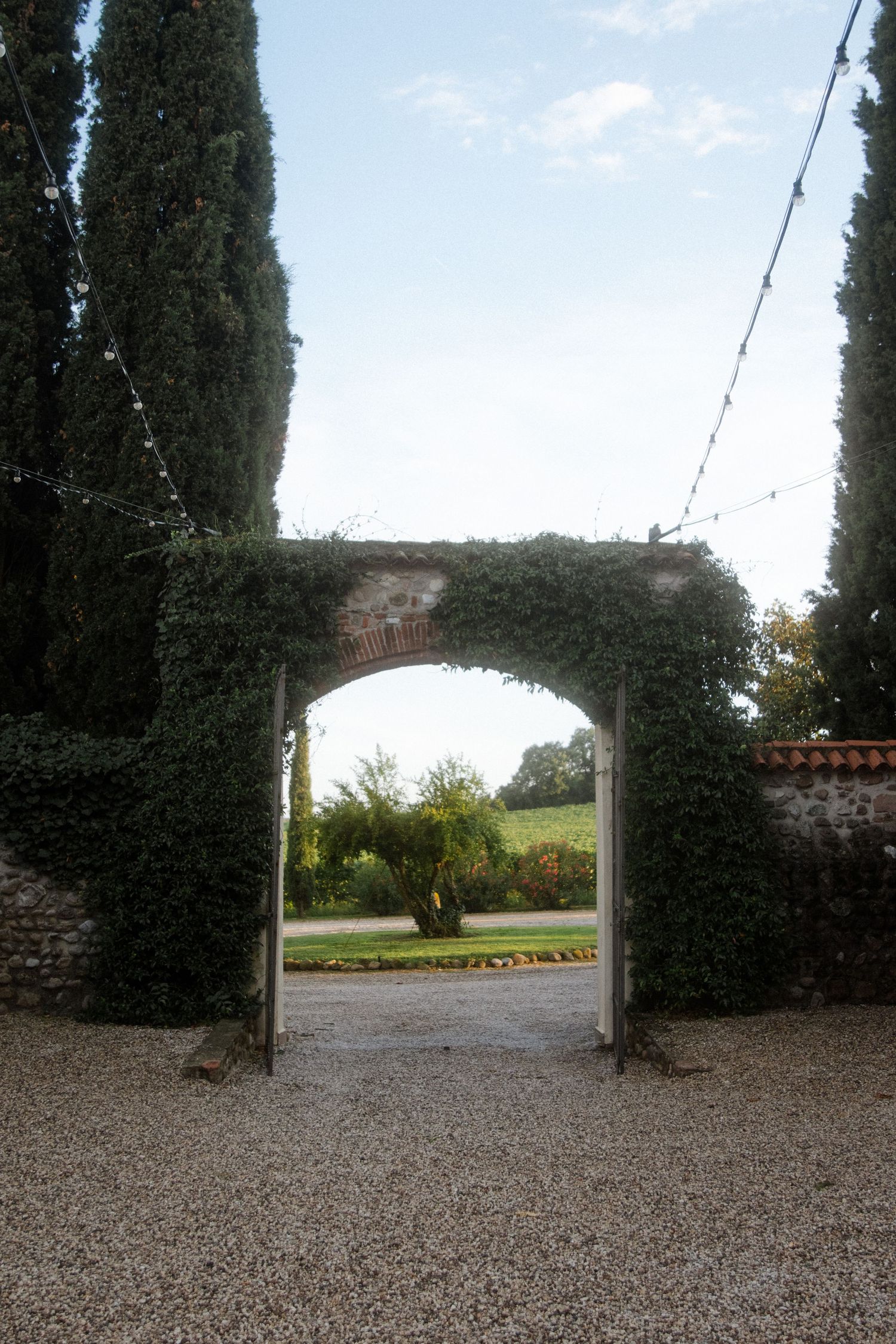 Ivy-covered stone archway entrance with cypress trees and string lights.