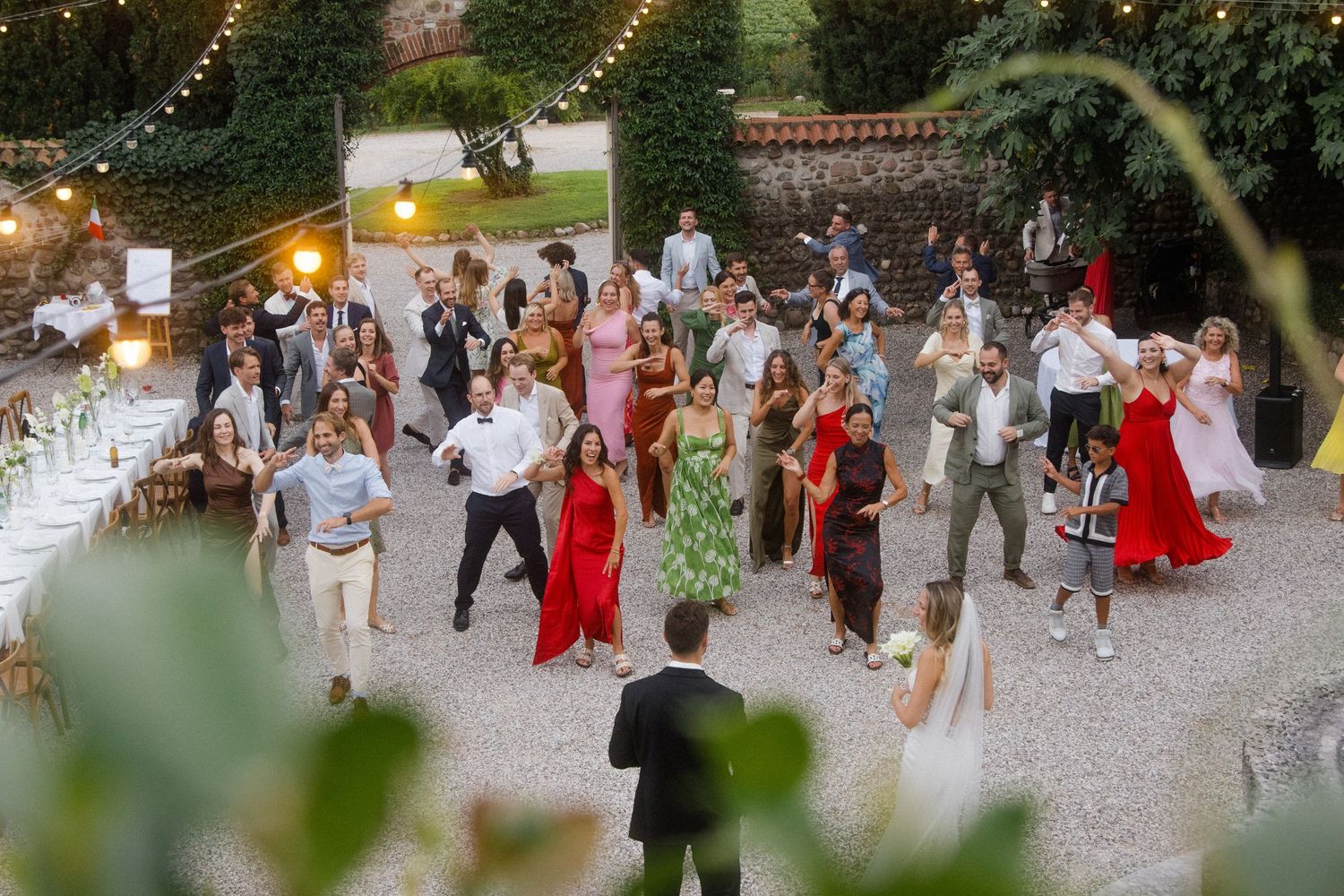 Outdoor wedding reception with guests dancing under string lights and greenery.